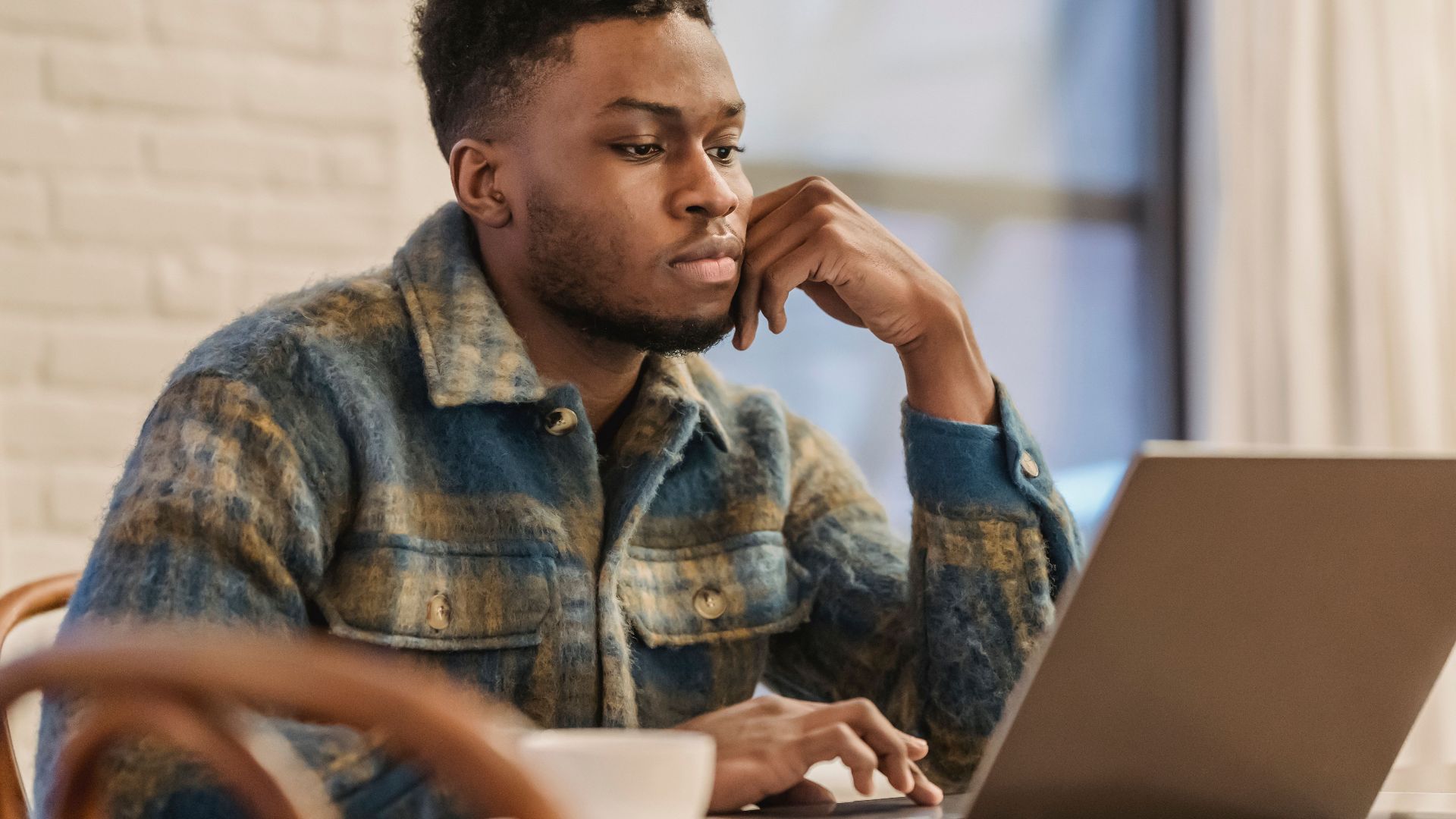 Concentrated bearded African American male freelancer in casual clothes sitting at table with laptop and leaning on hand while working on remote project in home workspace
