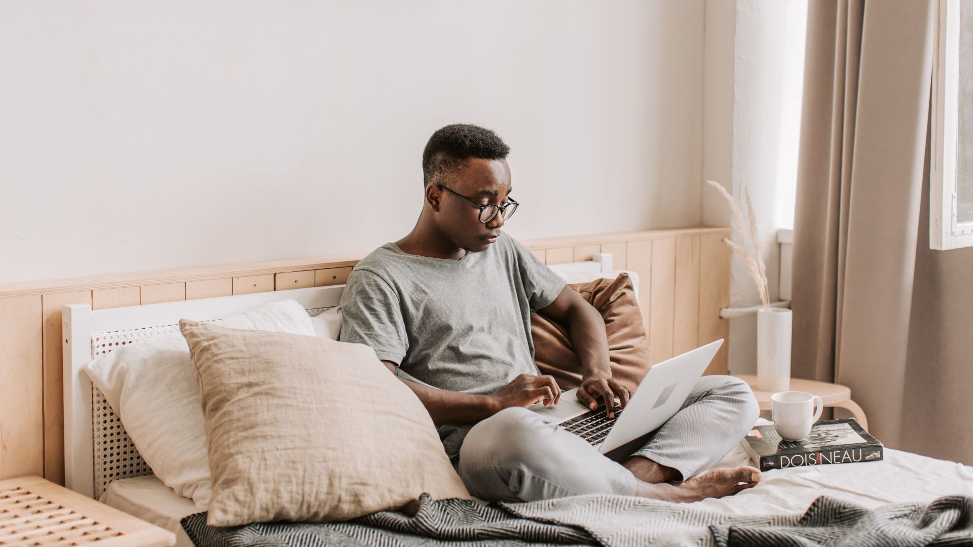 A man in casual attire working on his laptop from a comfy bed in a serene bedroom setting.