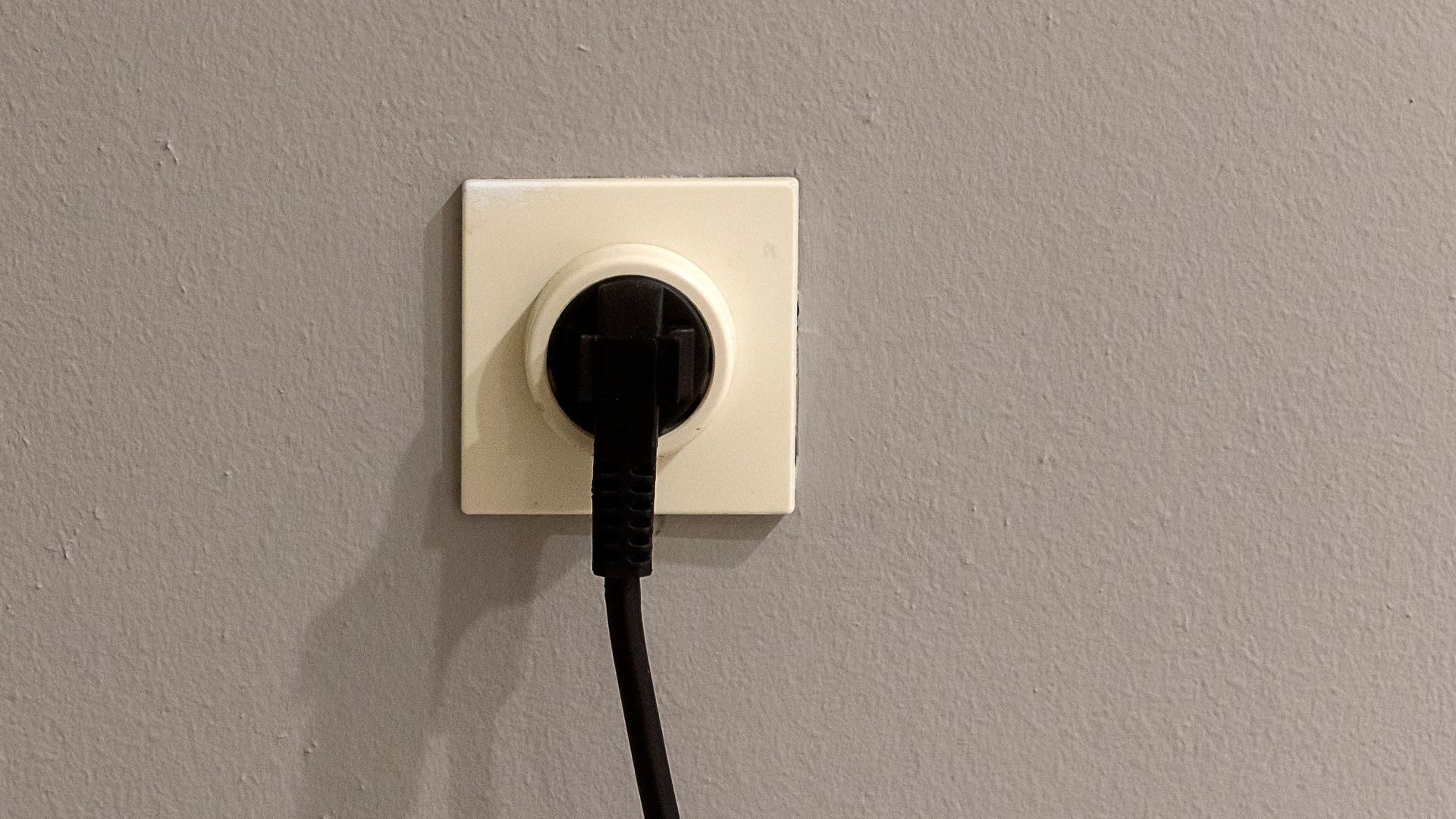 A black plug inserted into a white wall socket on a plain grey wall.