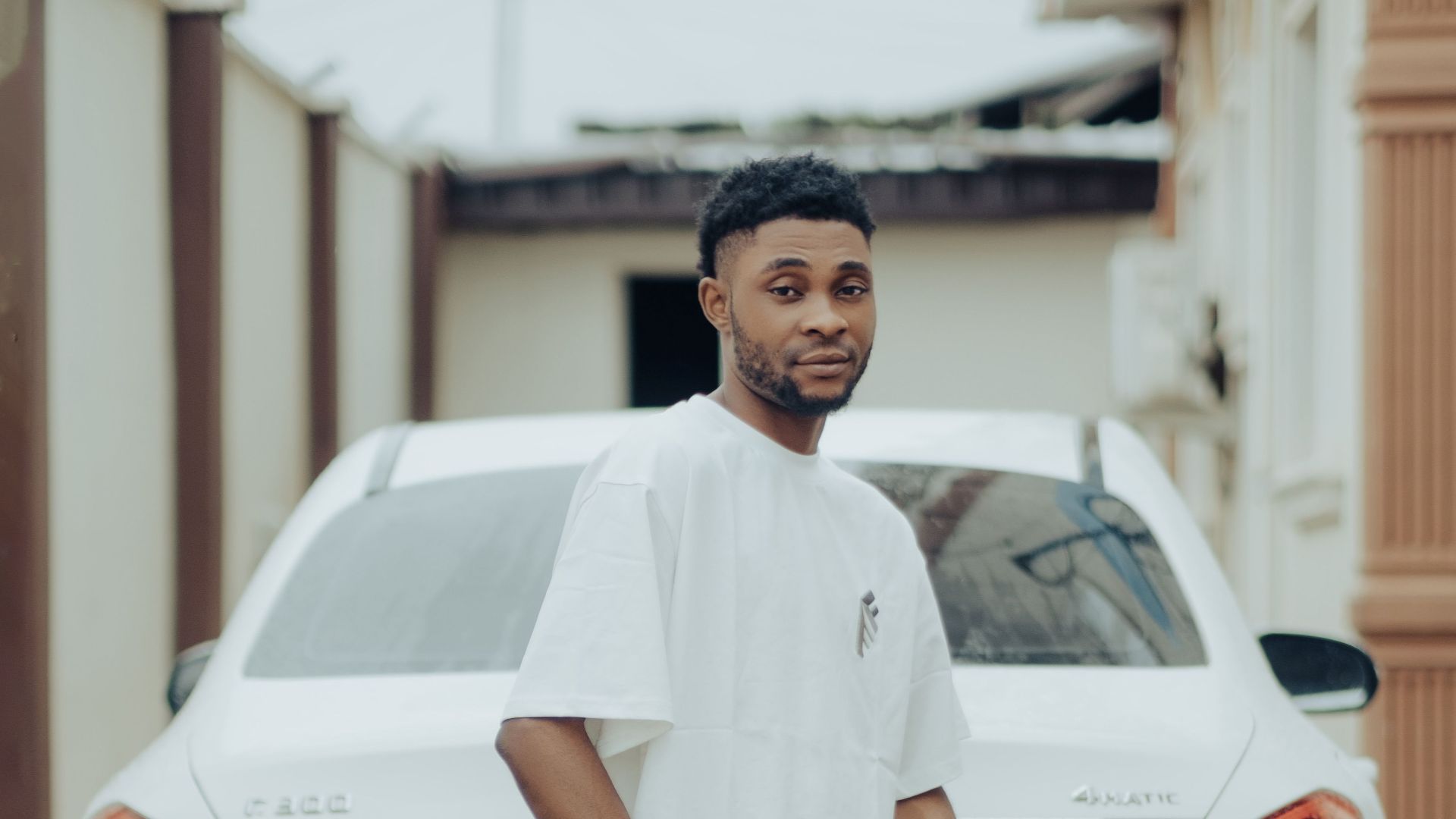 Young man in casual clothing standing by a white luxury car on a driveway.