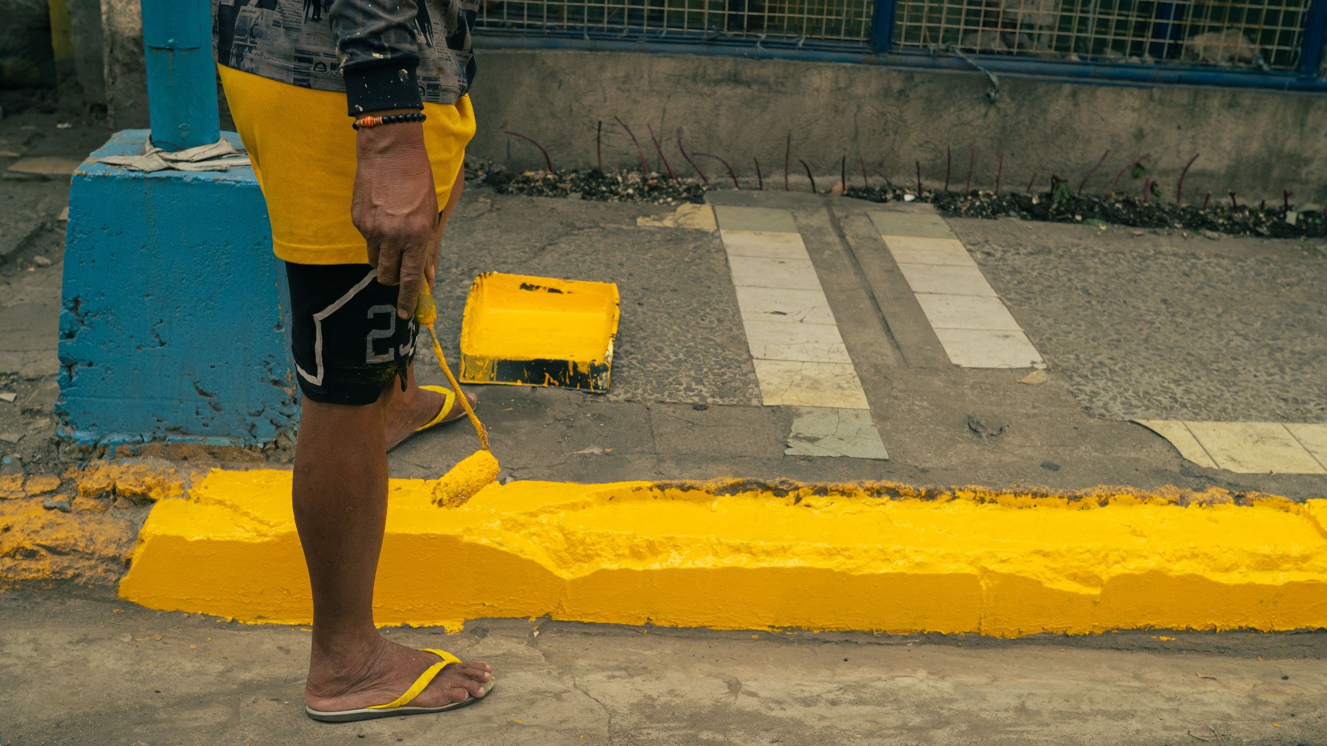 Street scene capturing a person in flip-flops painting a curb yellow with a roller.