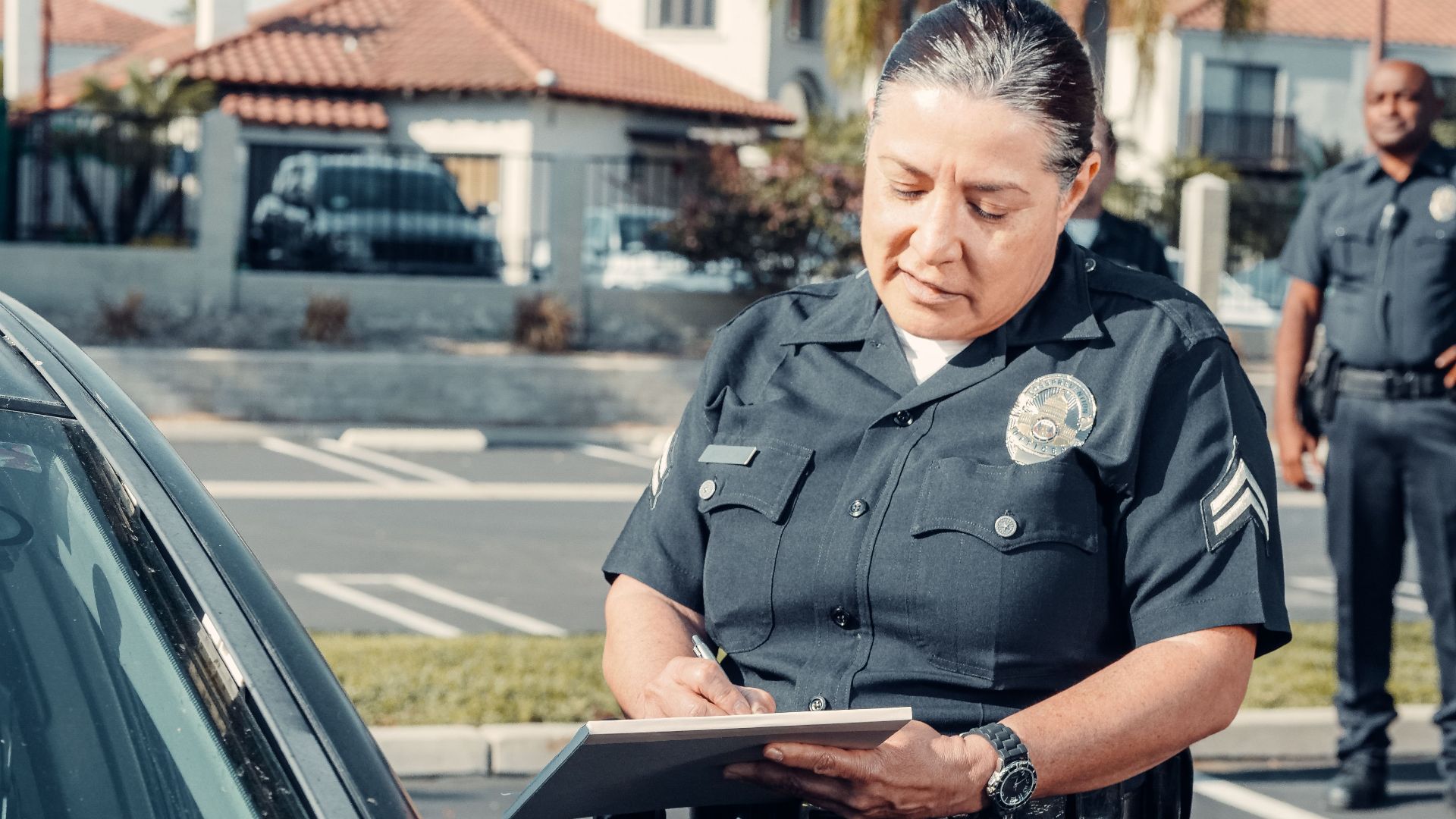 Police officer writing a ticket to a car in a sunny parking lot with a colleague in the background.