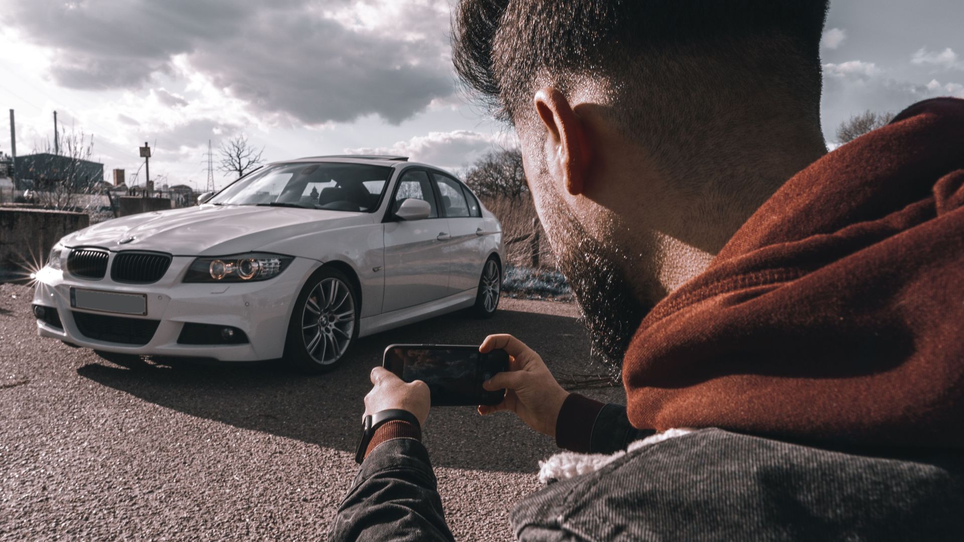 A man takes a picture of a white luxury car using a smartphone outdoors under a cloudy sky.