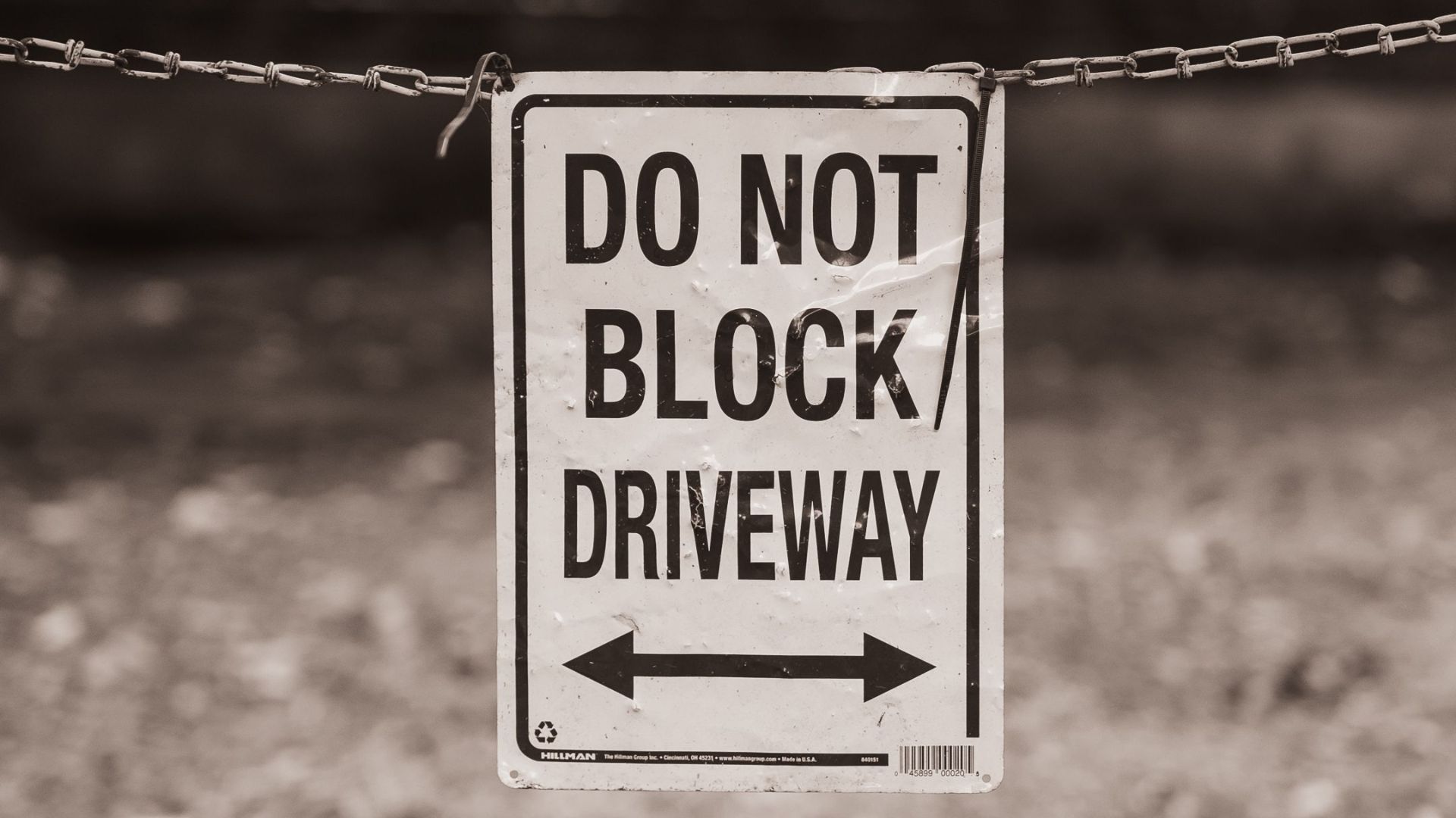 A vintage-style 'Do Not Block Driveway' sign hanging outdoors on a chain.