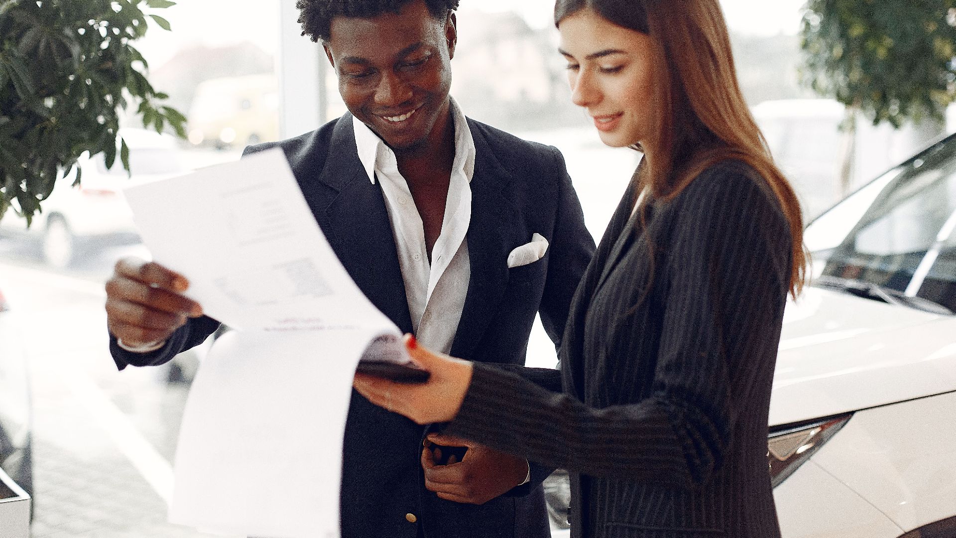 Cheerful smiling multiethnic businessman in classy suit and professional elegant female consultant standing close and reading contract details in car showroom