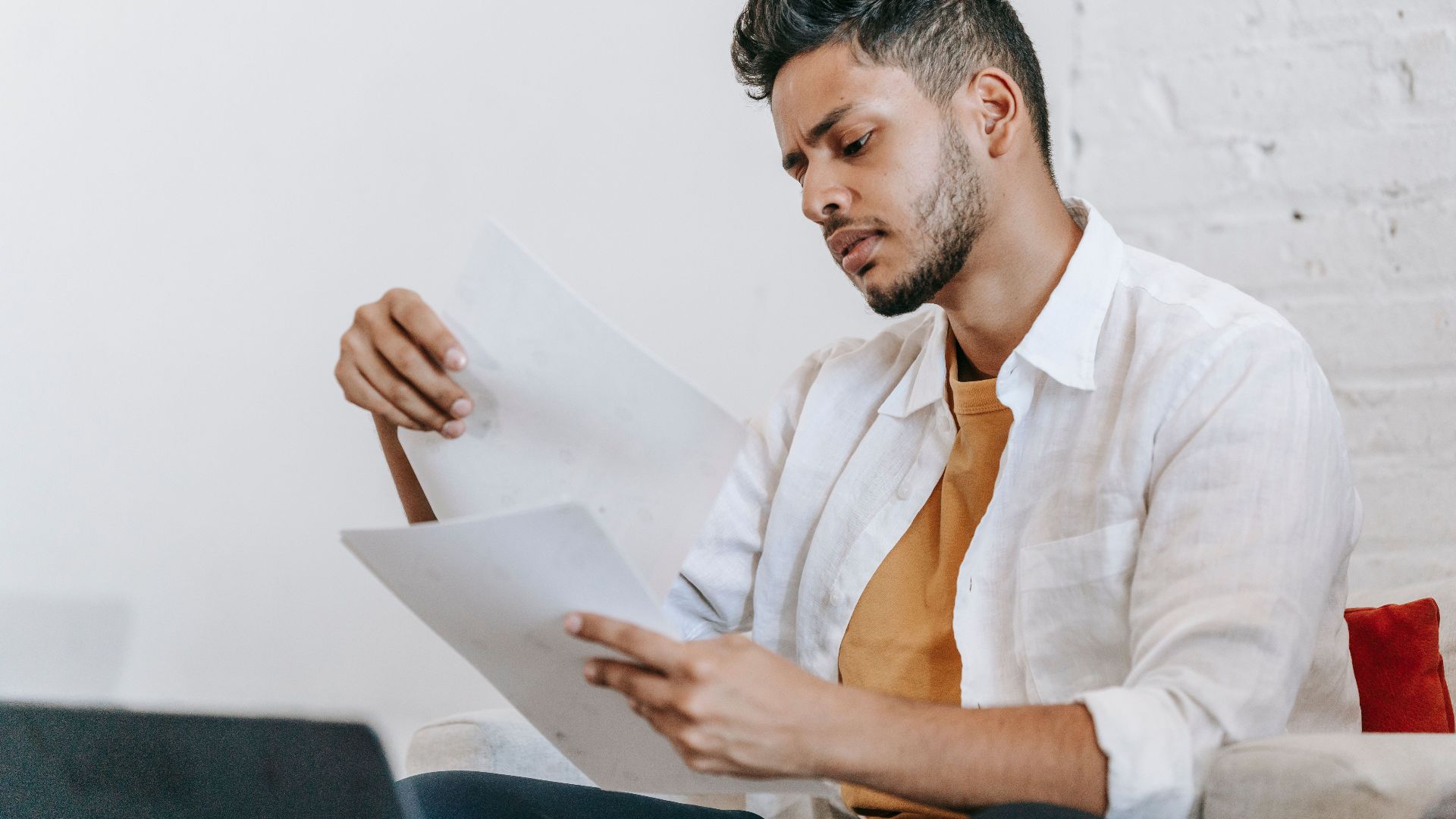 Concentrated ethnic male in casual wear reading documents and sitting on armchair near opened netbook