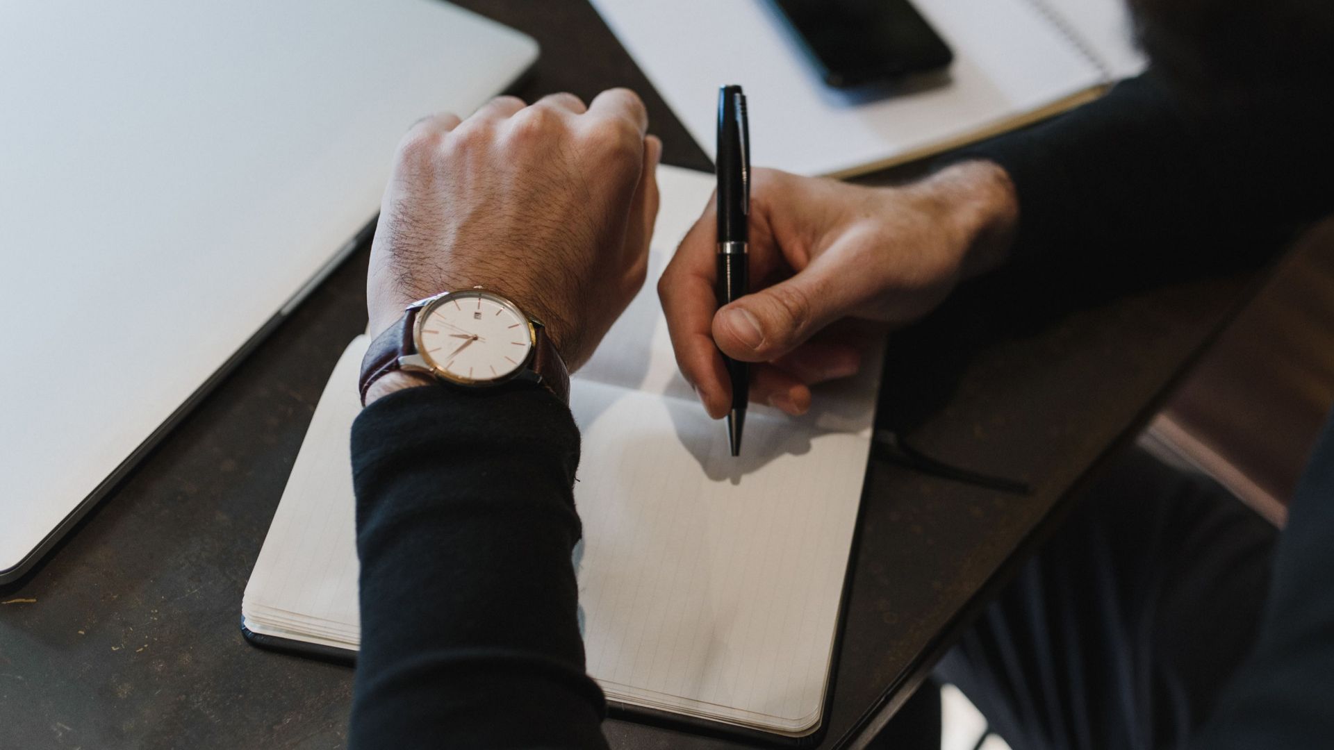 Close-up of a person writing in a notebook, wearing a watch, top view.