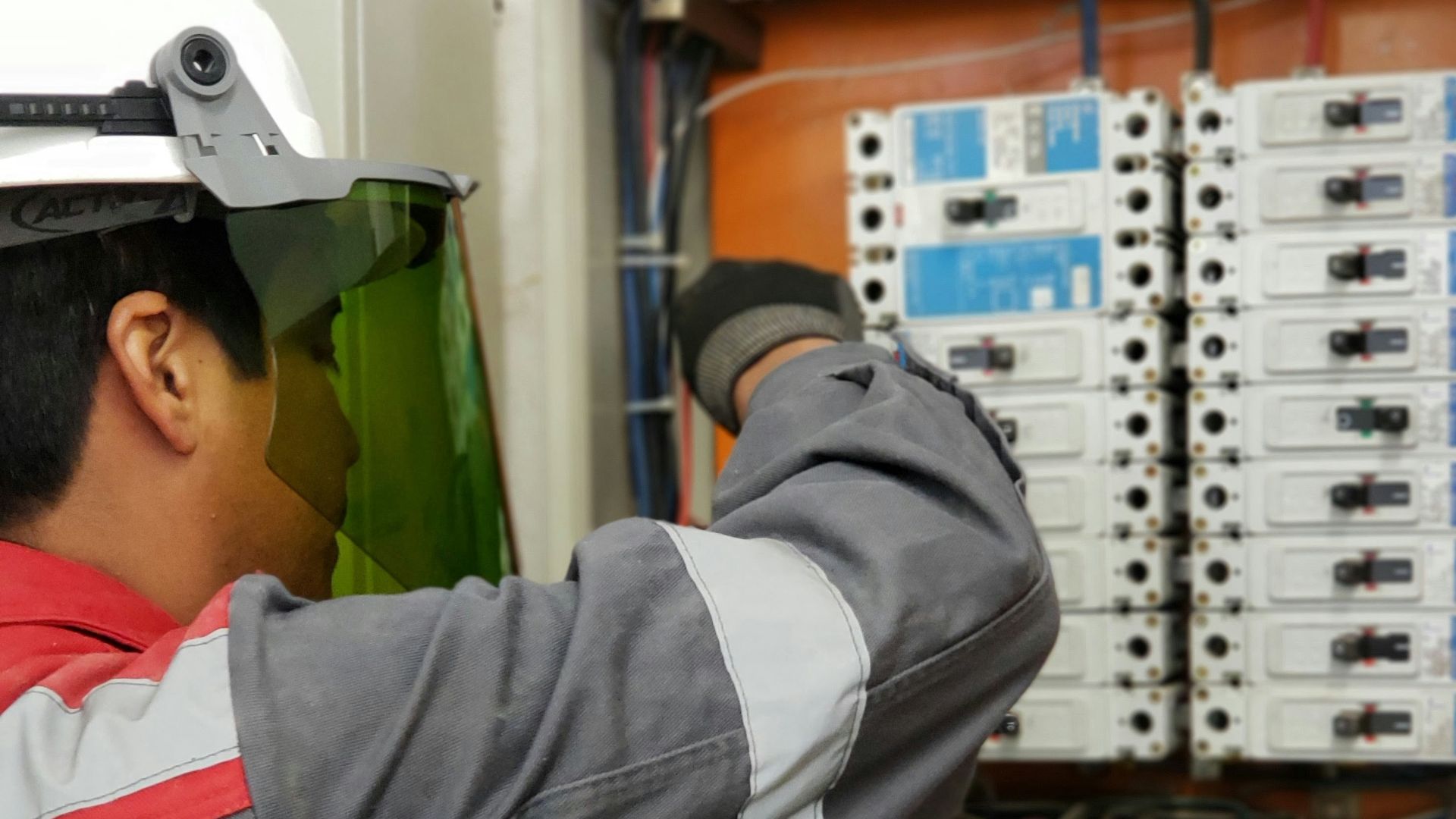 An electrician in a hard hat and face shield testing an electrical panel