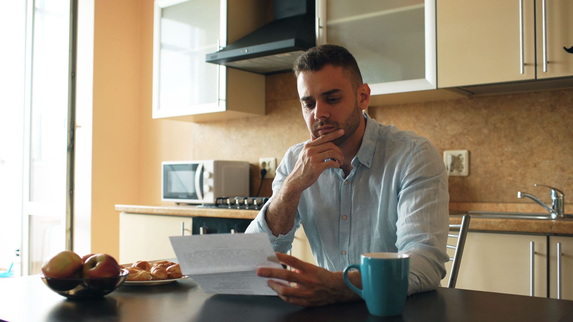 Thoughtful man reads a letter in a cozy kitchen setting with sunlight.