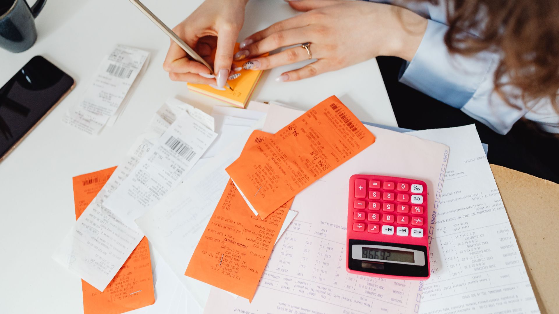 A woman writes down financial details using a calculator, receipts, and documents.