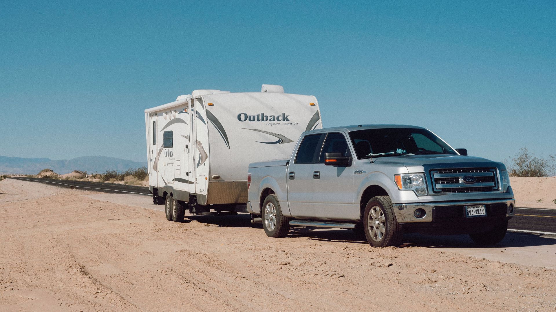 Silver truck towing an Outback trailer on a desert road under clear blue skies.