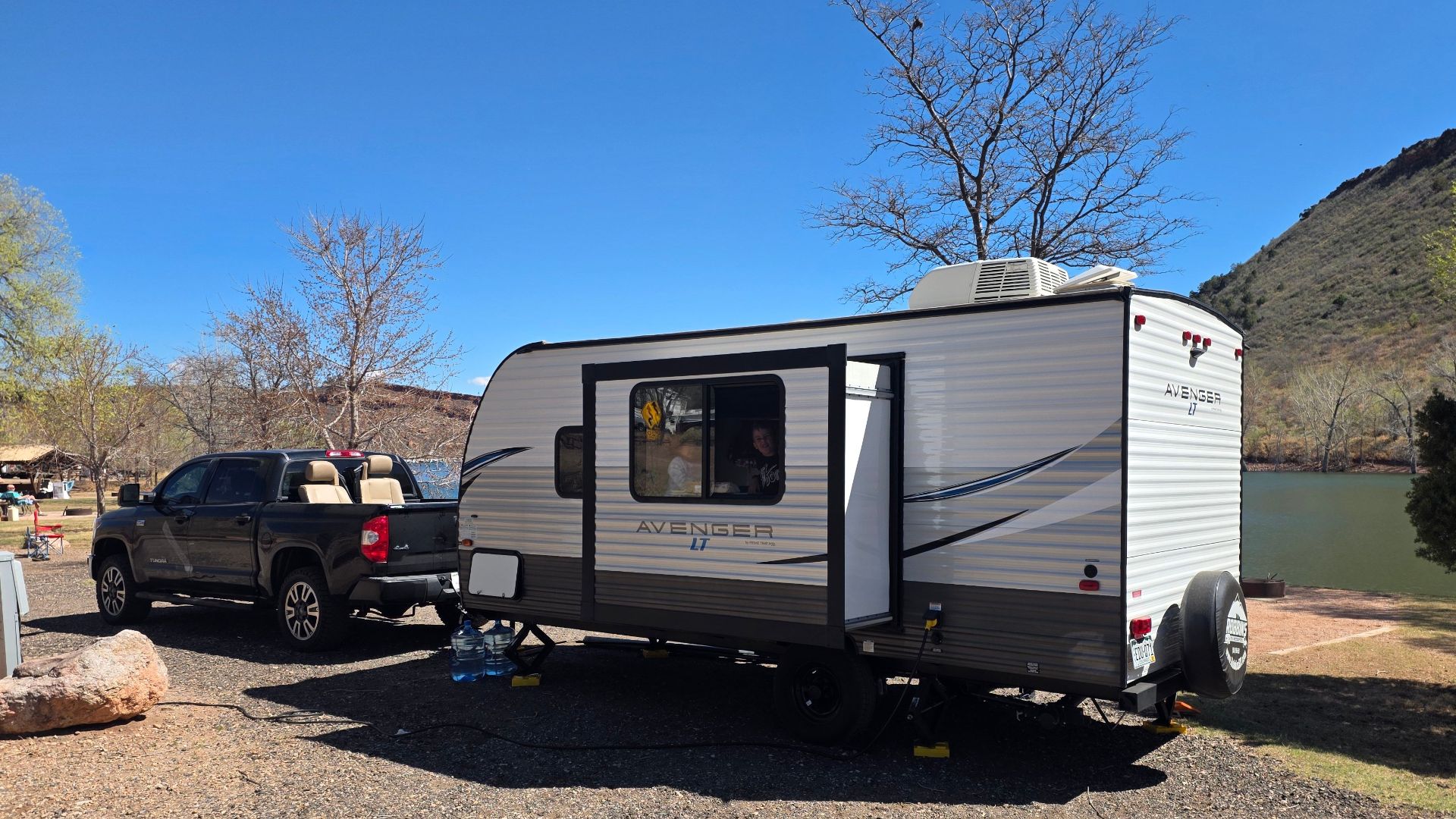 A scenic daytime camping setup with a caravan and truck by a lake in a natural setting.