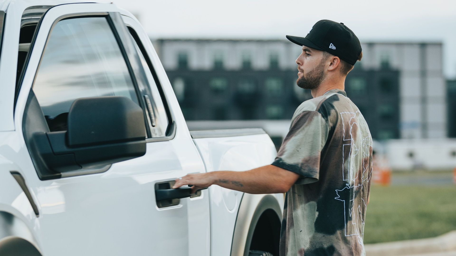 A man in casual attire opens the door of a parked white pickup truck outside.