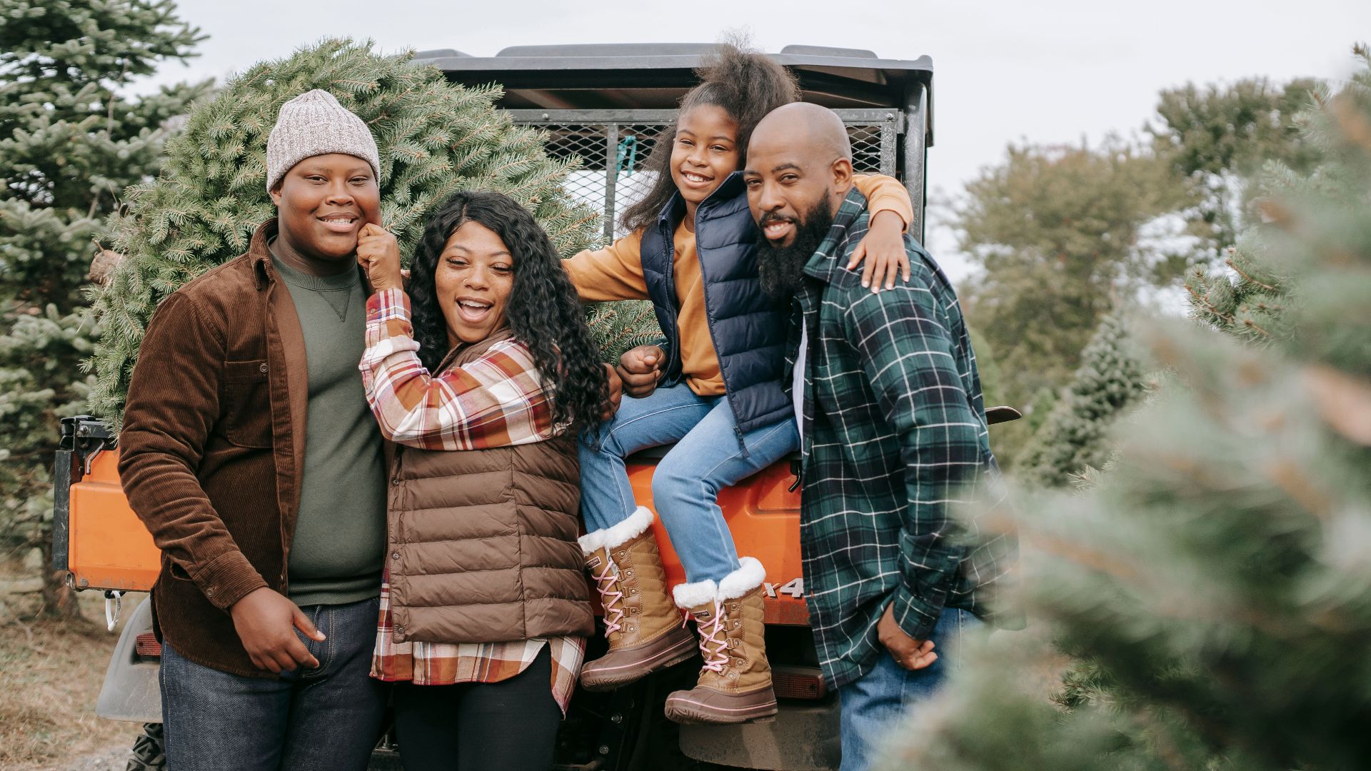 Cheerful African American parents embracing daughter and son near transport and coniferous trees while looking at camera in countryside