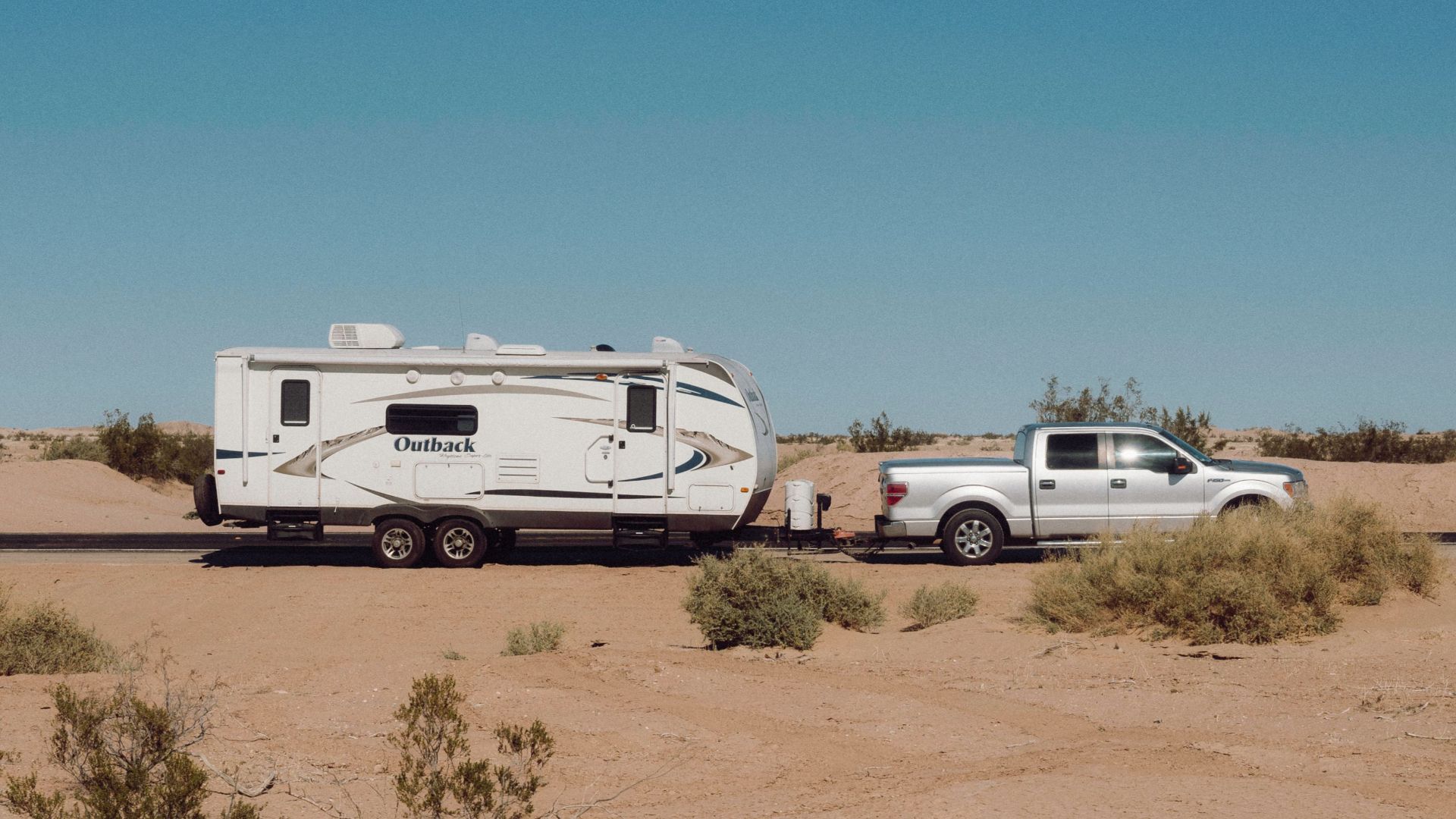A camper trailer towed by a truck travels through a remote desert landscape under a clear blue sky.