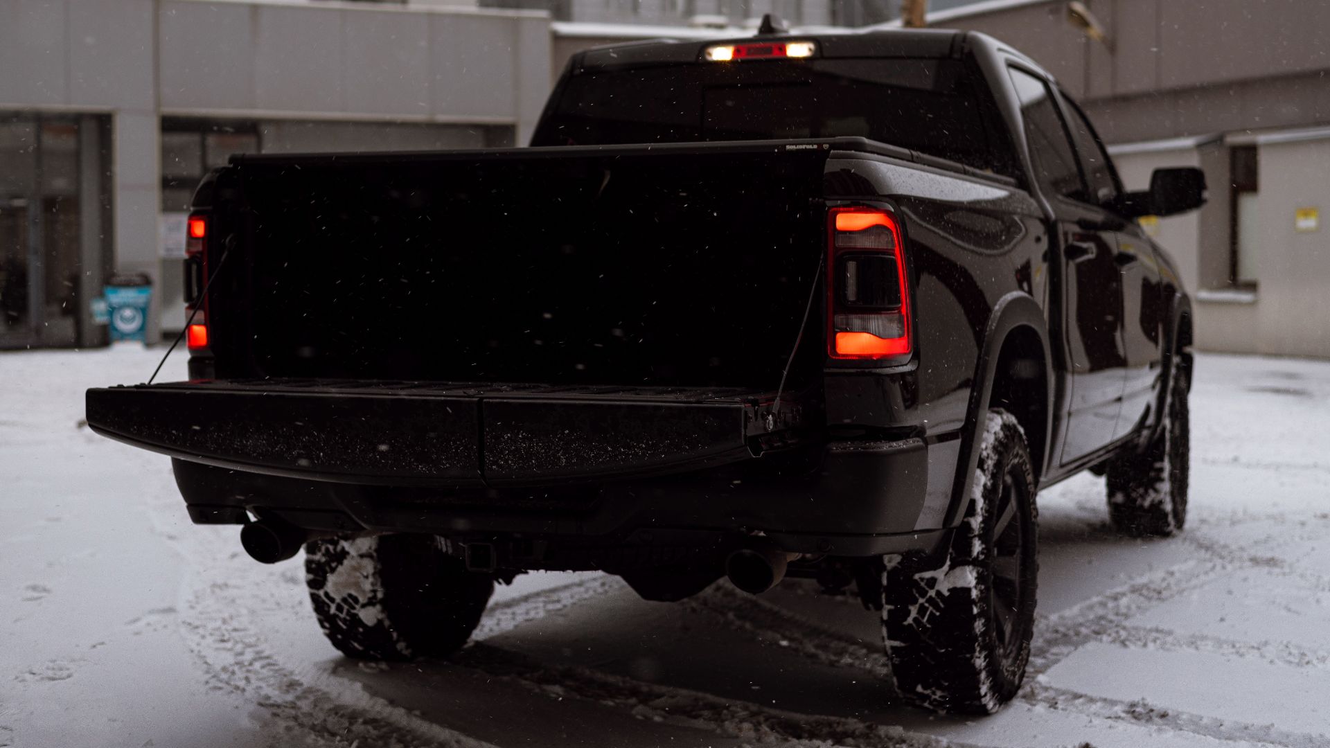 Black pickup truck parked on snow-covered urban street with falling snowflakes.