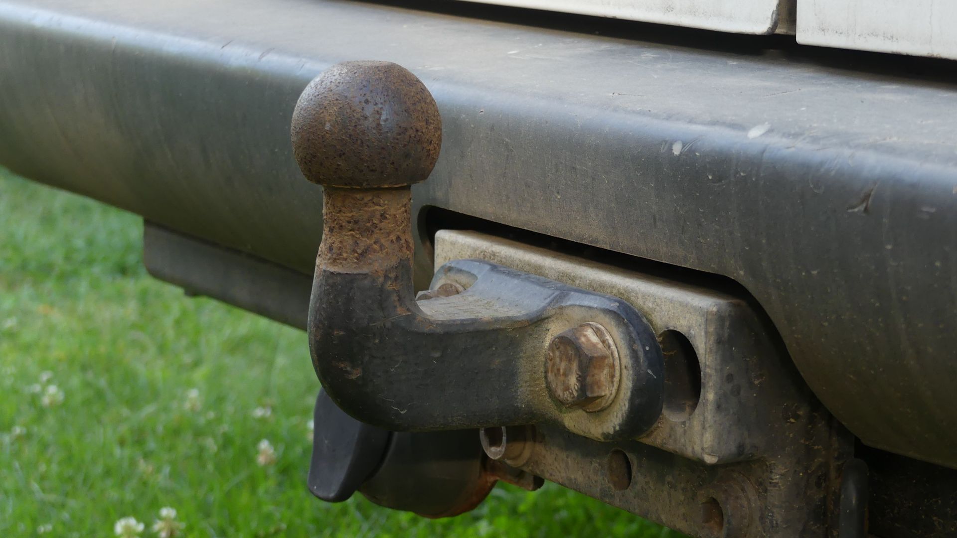 Close-up of a rusty tow hitch on the rear bumper of a vehicle against a grassy background.