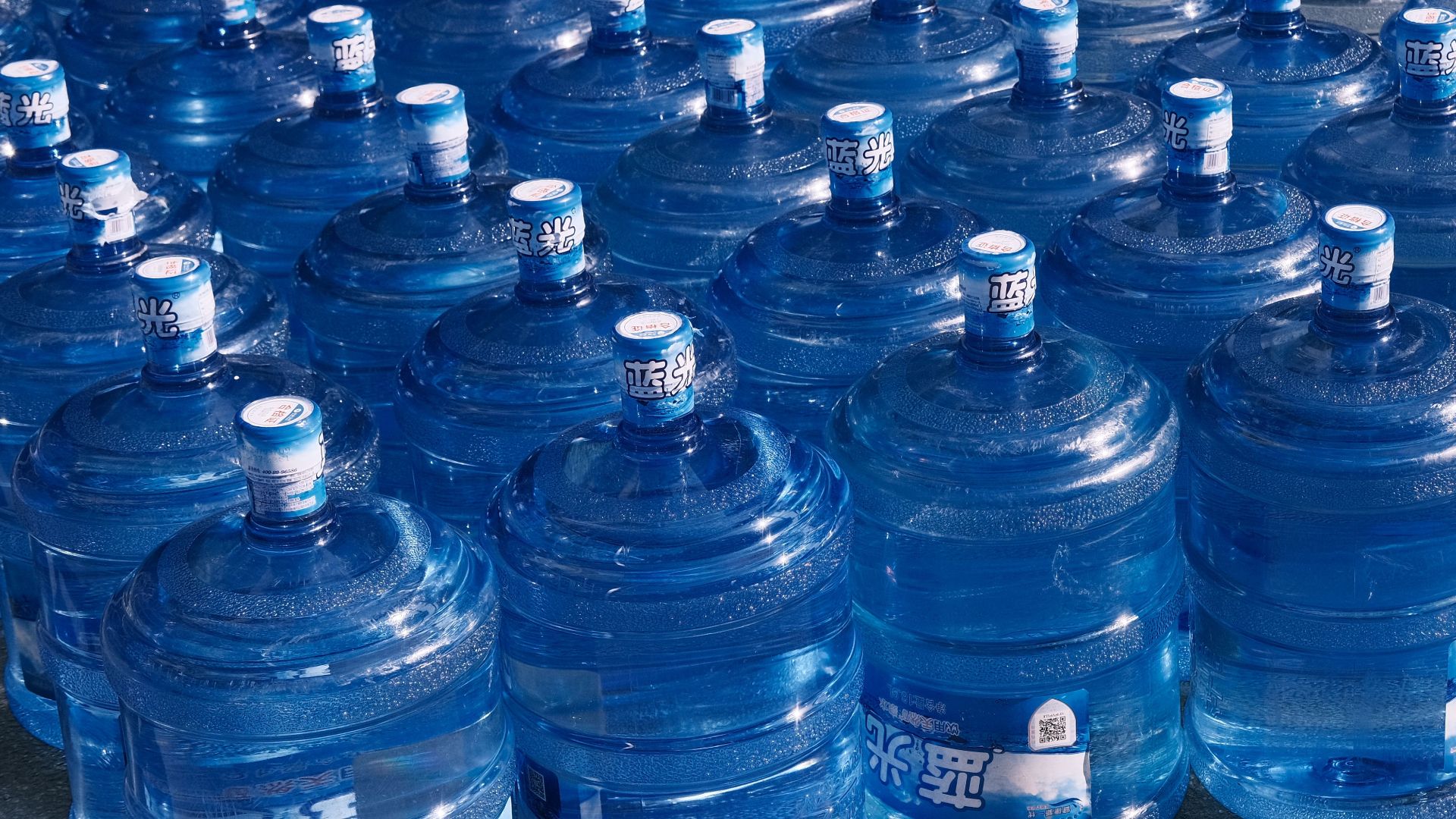 A collection of blue plastic water jugs lined up on an urban street in sunlight.