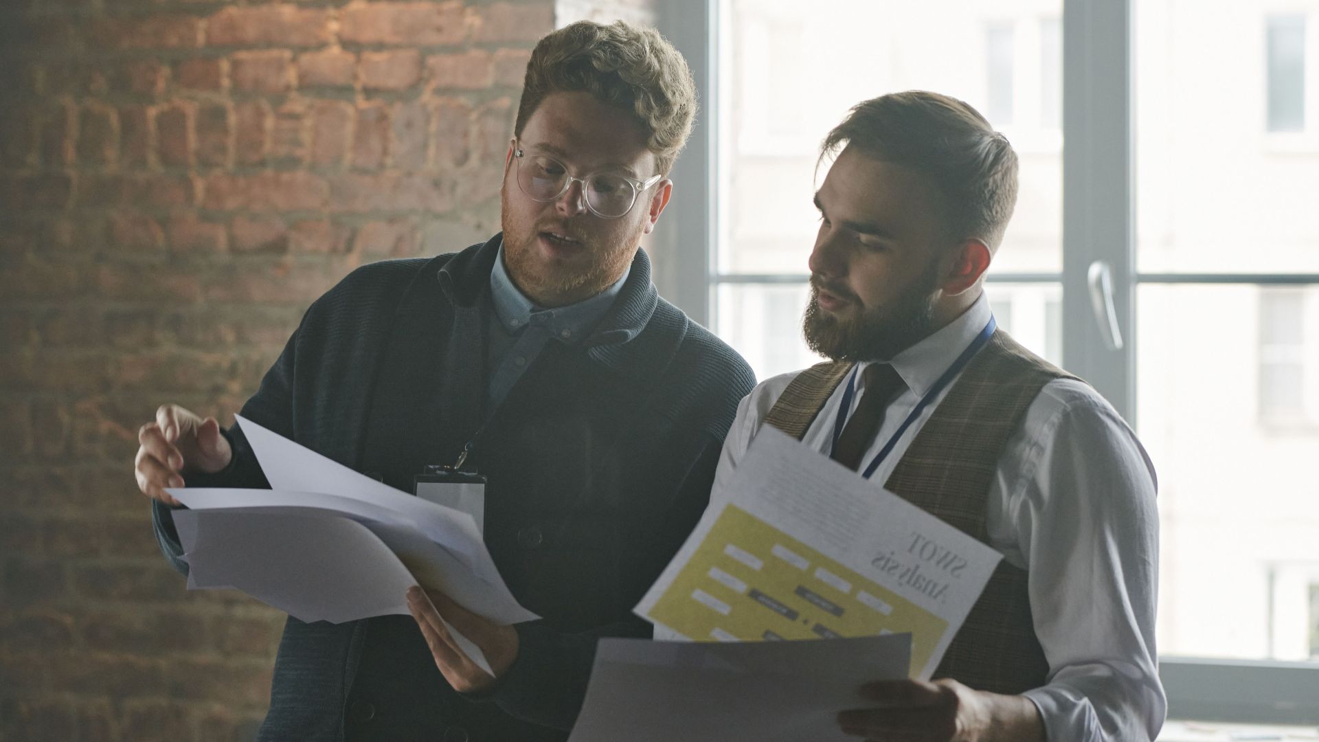 Two businessmen discussing documents in an industrial-style office setting.