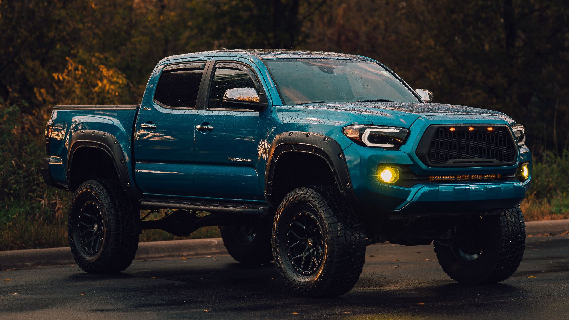 A lifted blue pickup truck parked on a wet road in a wooded environment.