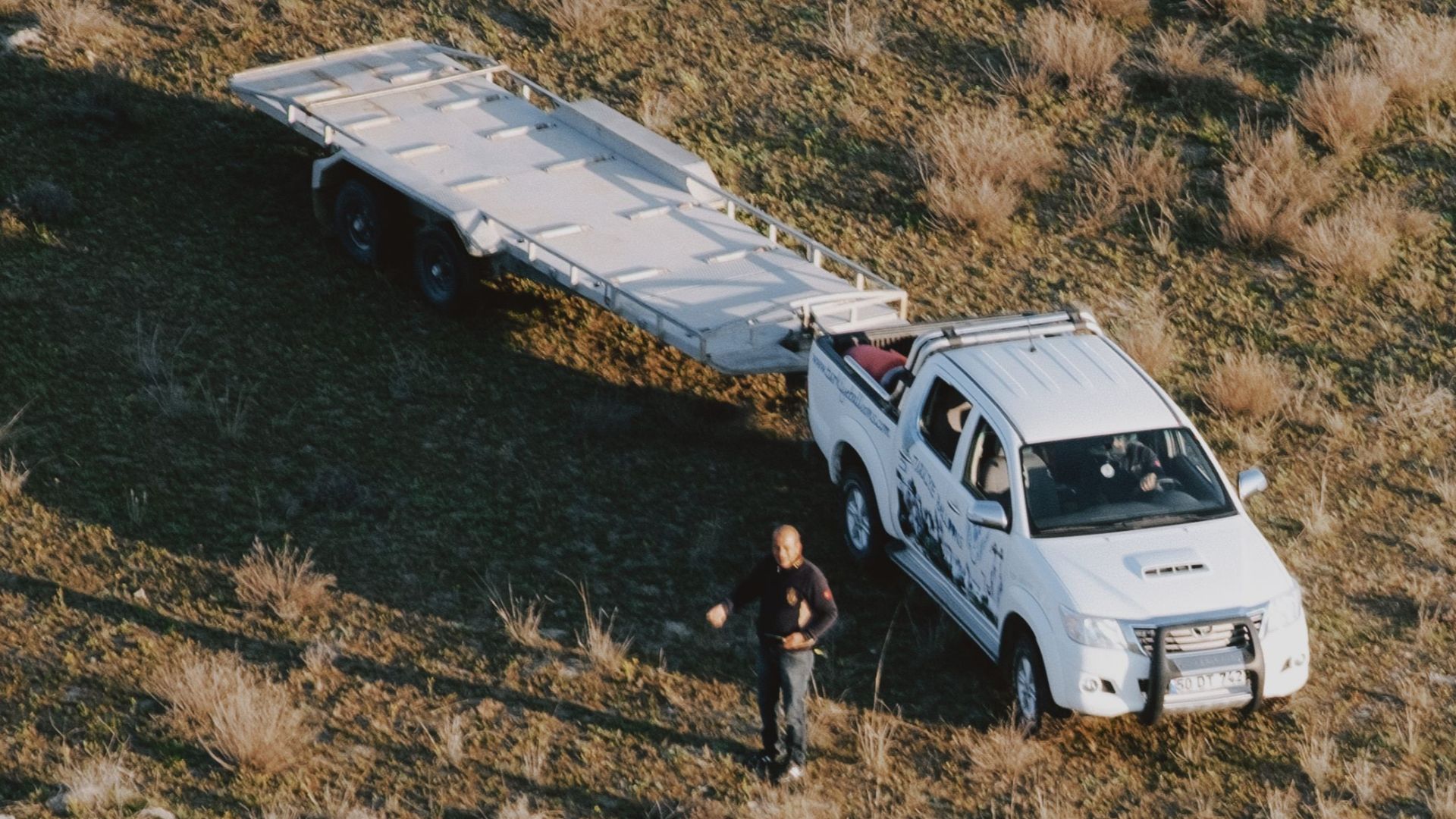 Overhead shot of a white truck with a trailer on a dry, grassy landscape. High contrast daylight scene.