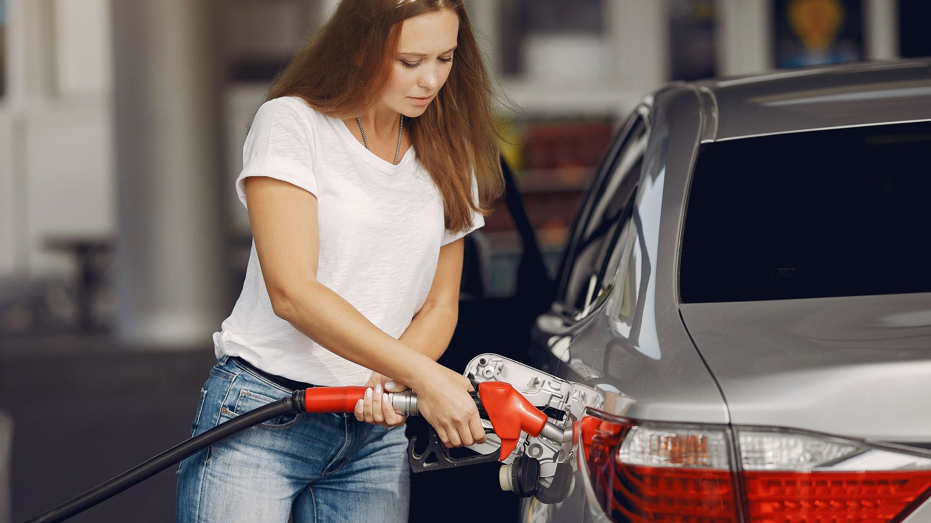 Attentive female driver in casual outfit and headband filling up modern automobile with automotive fuel gun on petrol station while looking down