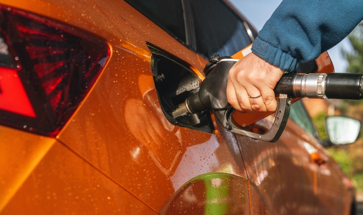 Refueling an orange car at gas station during daylight hours in an urban area with visible greenery in the background