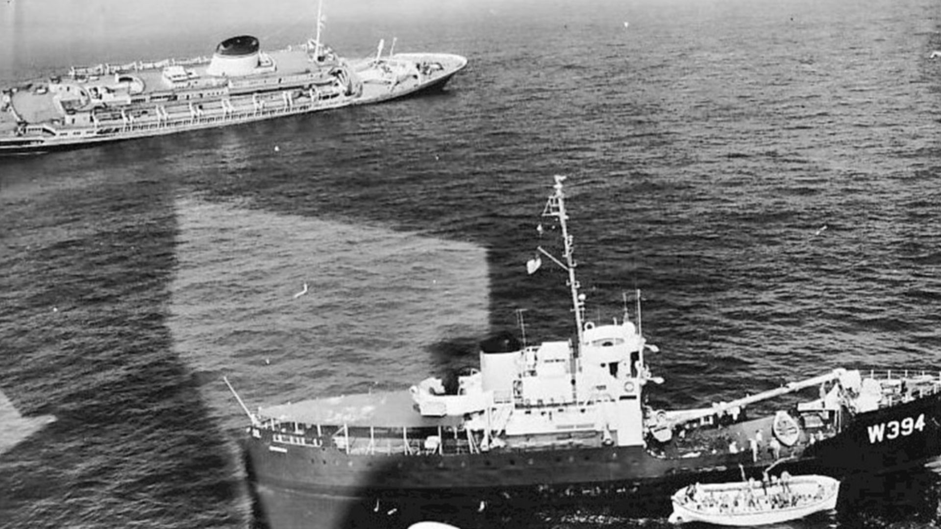 Photo of a  lifeboat of passengers from the sinking liner Andrea Doria being rescued by another vessel.  Photo identifies the craft as possibly being a buoy tender. Andrea Doria is seen in the background listing to the right--this is the dise where the co