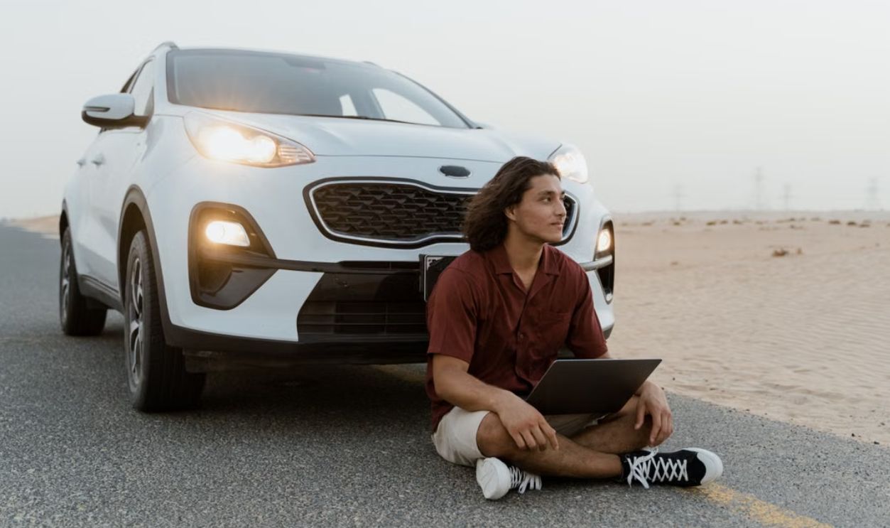 A man with a laptop sitting by an SUV on a desert road, working remotely.