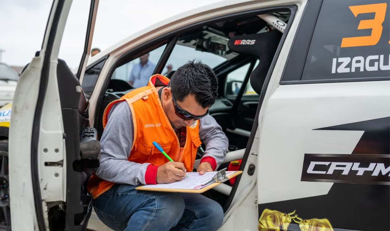 A motorsport official reviews documents while sitting by a rally car during a race event.