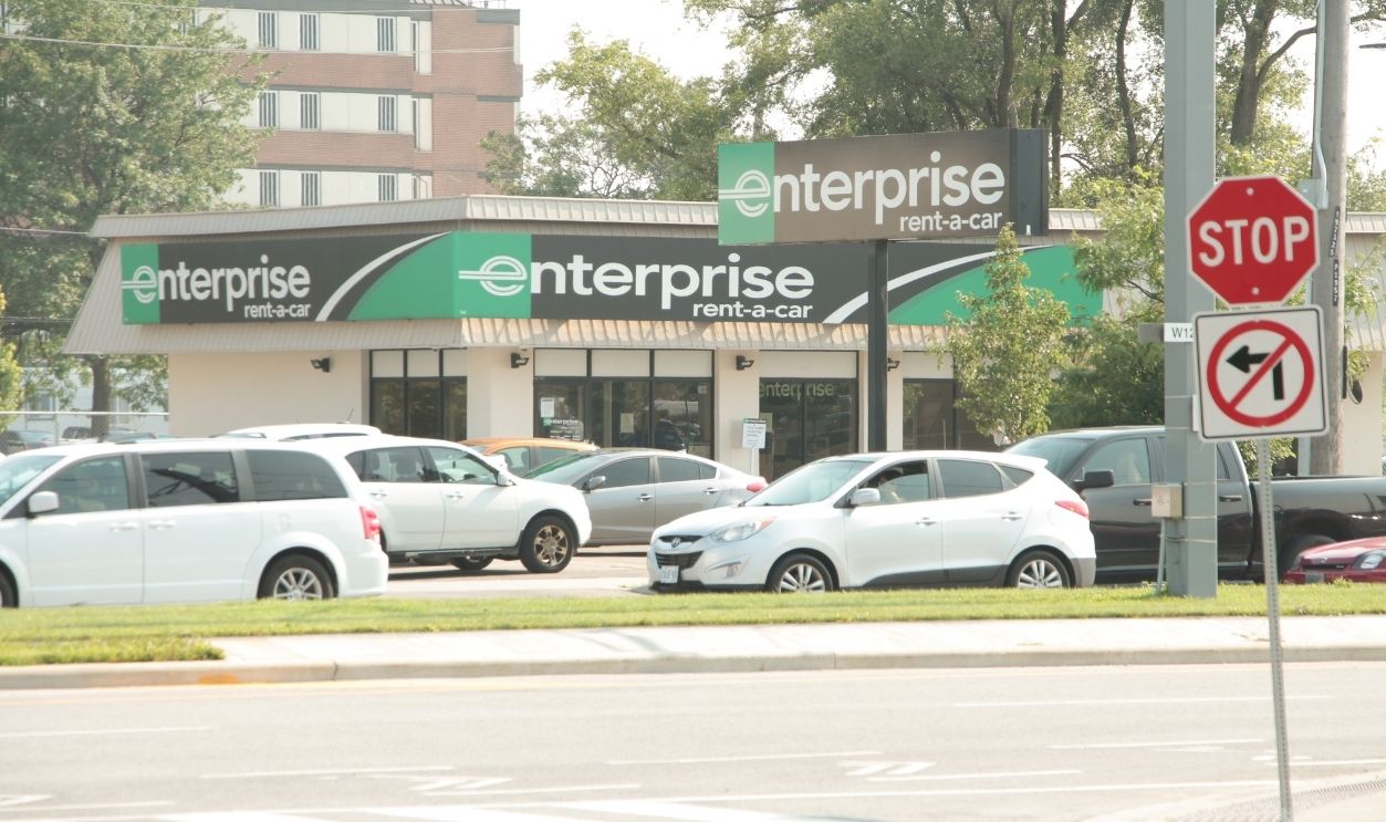 tor, canada - august 16, 2023: enterprise rent-a-car store storefront green black with sign logo shot from across the street road with cars traffic vehicles passing in front