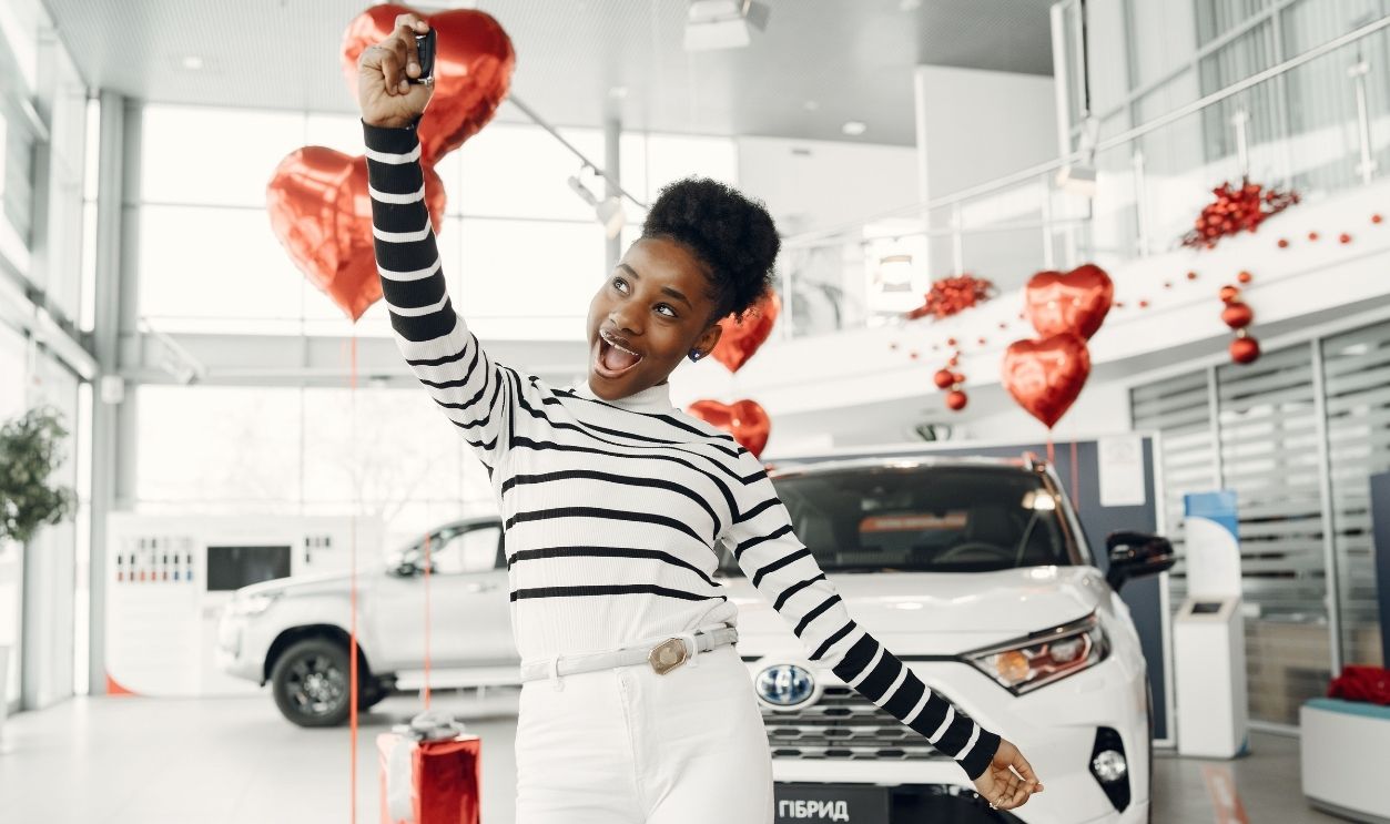 Woman excitedly holds car key in dealership with red balloons, symbolizing celebration and achievement