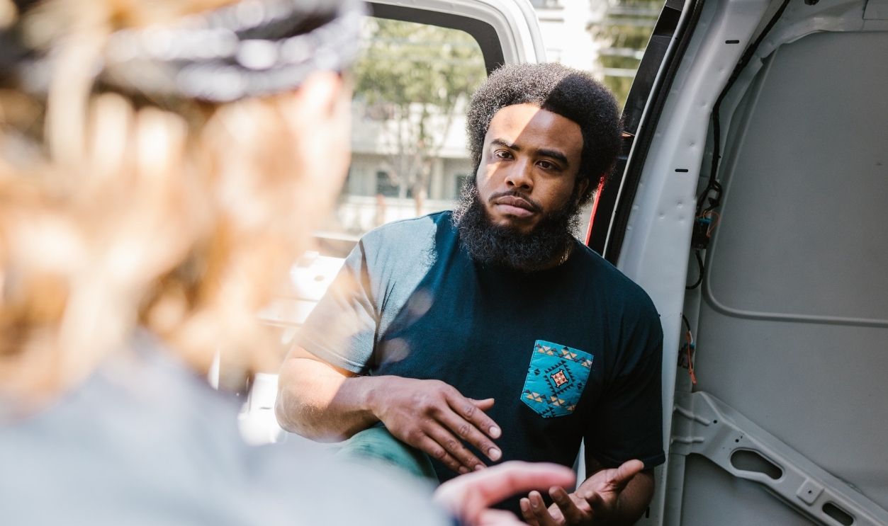 Two men engage in conversation next to a parked van on a sunny day.