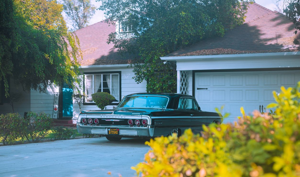 A classic black Chevrolet Impala parked in a suburban driveway surrounded by greenery