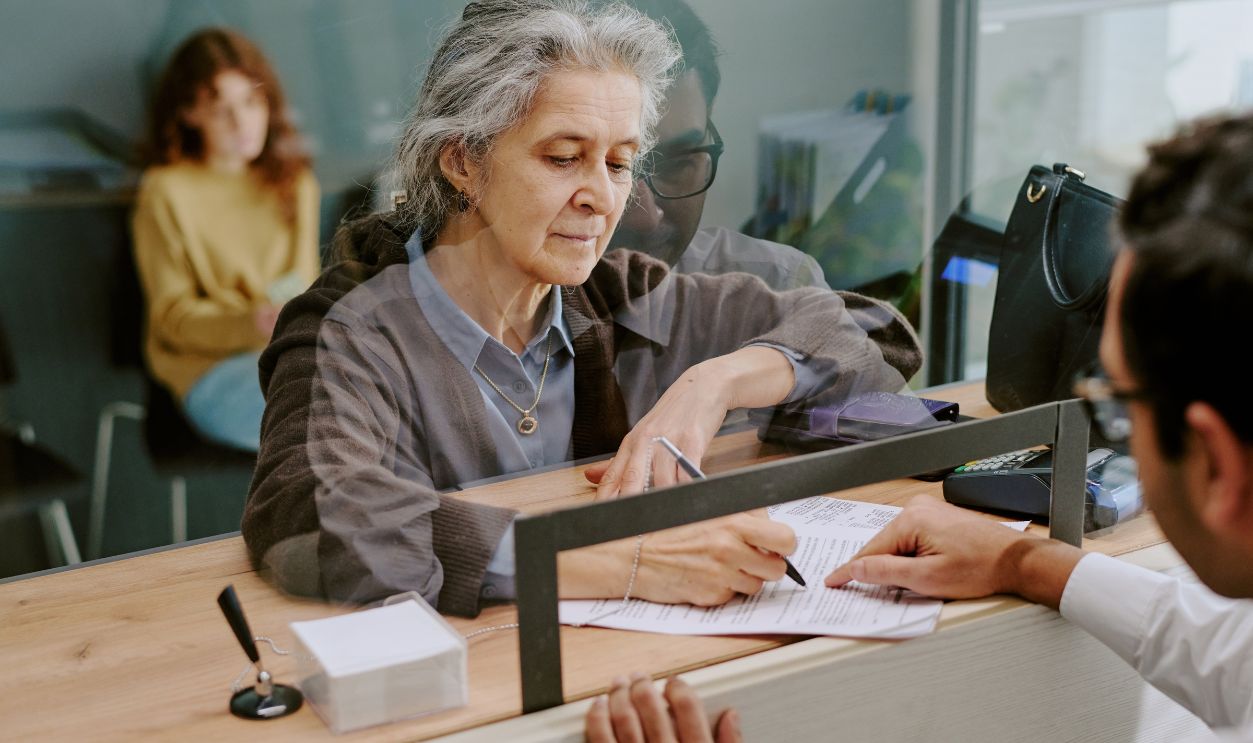 Elderly woman signing official documents at banking counter with teller assisting, seated in modern office setting with reflective glass partition