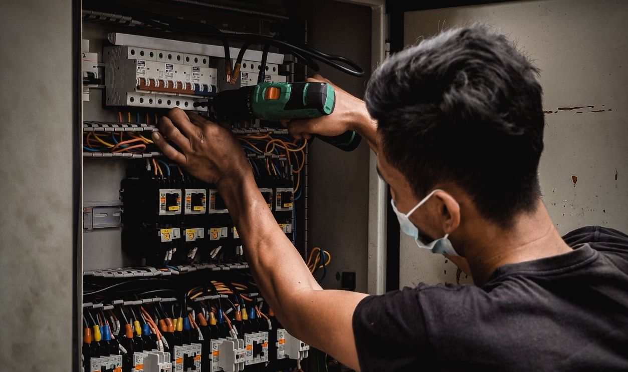 Technician working on circuit breaker panel, adjusting electrical wires indoors.