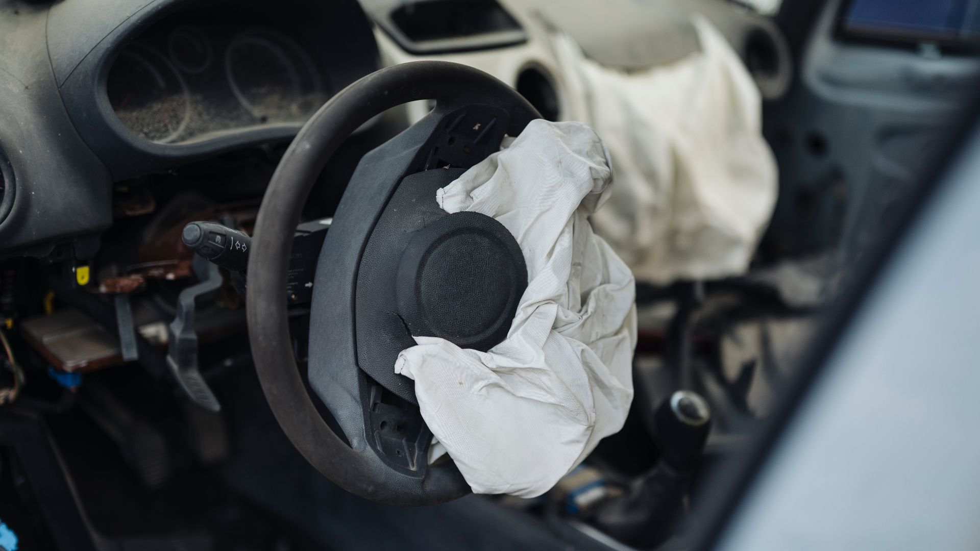 An abandoned car interior shows signs of neglect. The steering wheel displays a deployed airbag, and dust covers the surfaces. The setting appears to be a forgotten area with little activity.