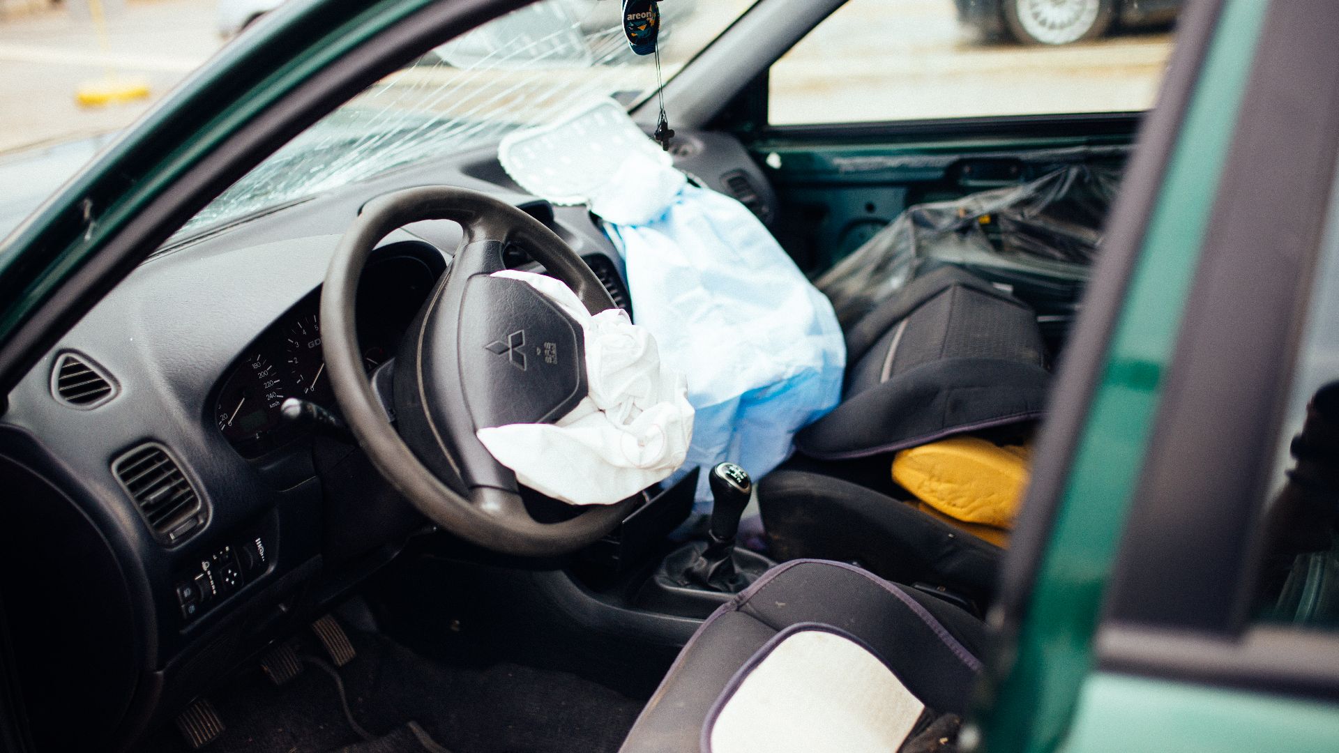 The car interior shows severe damage from a crash, with the airbag deployed and debris scattered. The accident took place on a busy urban street during daylight hours.