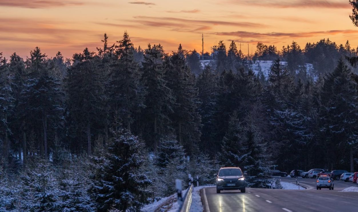 A car drives along a snowy road through conifer forests at sunset, creating a serene winter scene