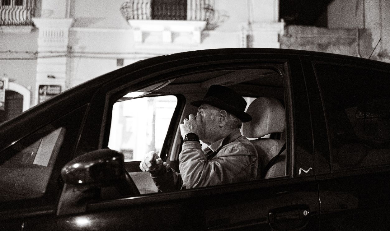 Black and white image of an elderly man driving a car at night, captured in an urban setting