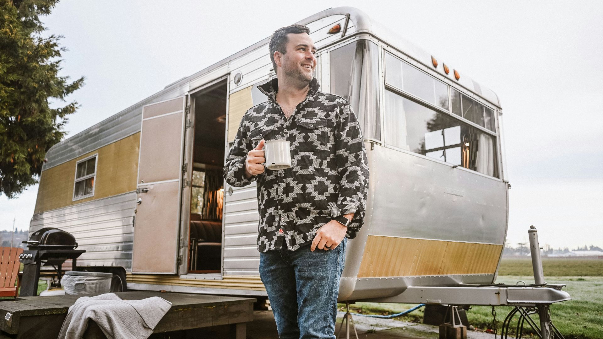 A man holding a mug stands outside a vintage camper