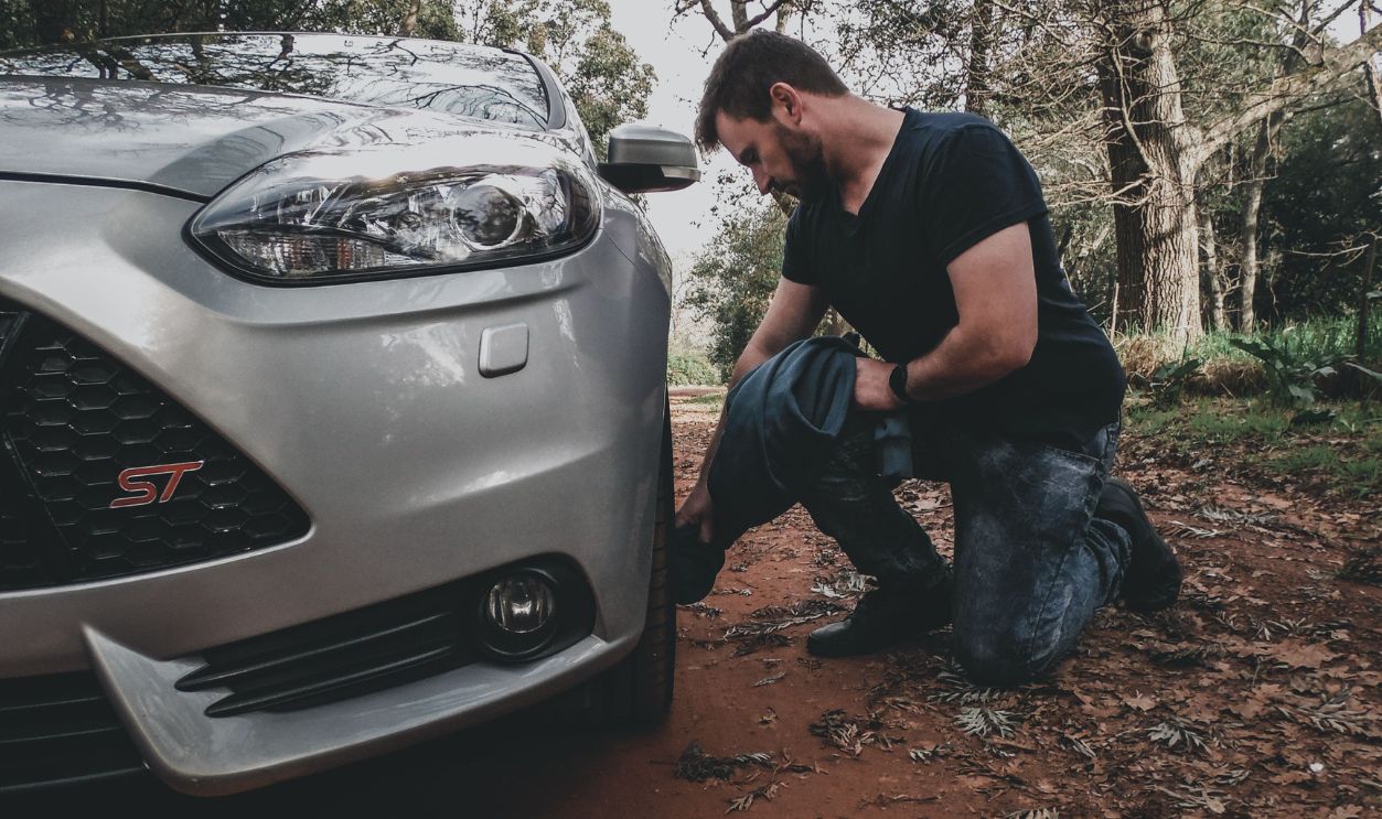 A man kneels to check the tire of a silver car in a forest