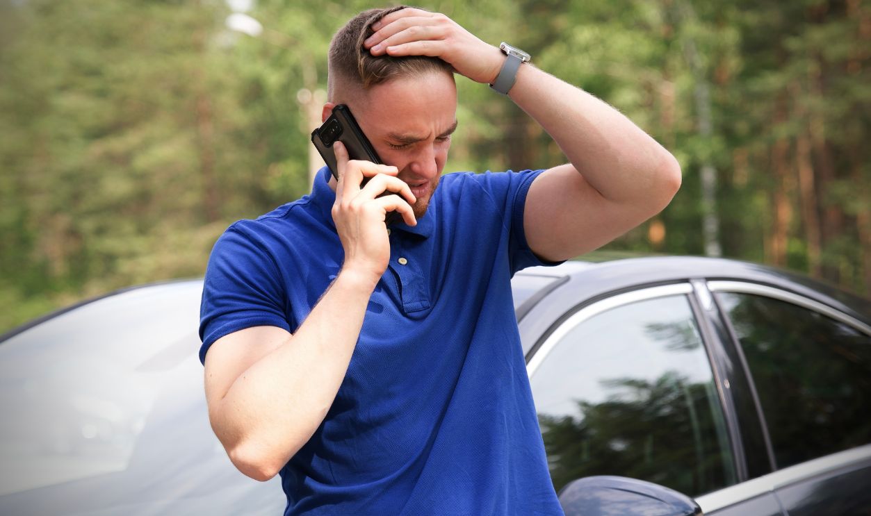 Shutterstock - 2182976321 - Sad upset guy driver is standing near car, calling emergency on cell mobile phone talking on smartphone. Young desperate man got into accident holding head with hand.