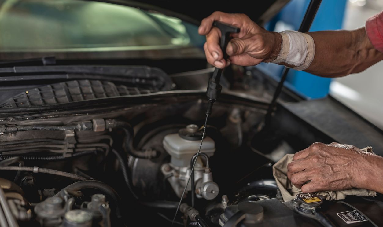 An old mechanic with an injured wrist checks the transmission fluid of an old sedan with a dipstick.