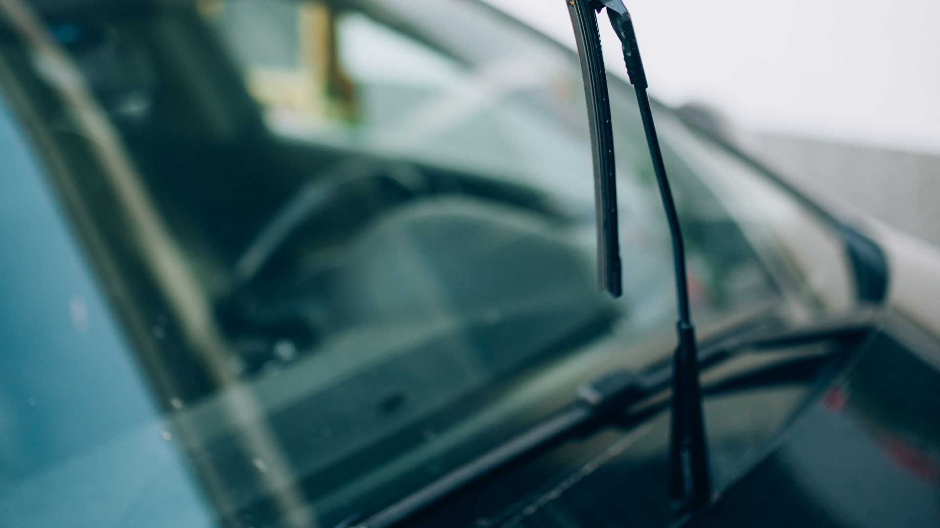 A car's windshield wiper stands upright while parked at a service station on a sunny day. The background features greenery and flower planters, emphasizing a calm suburban setting.