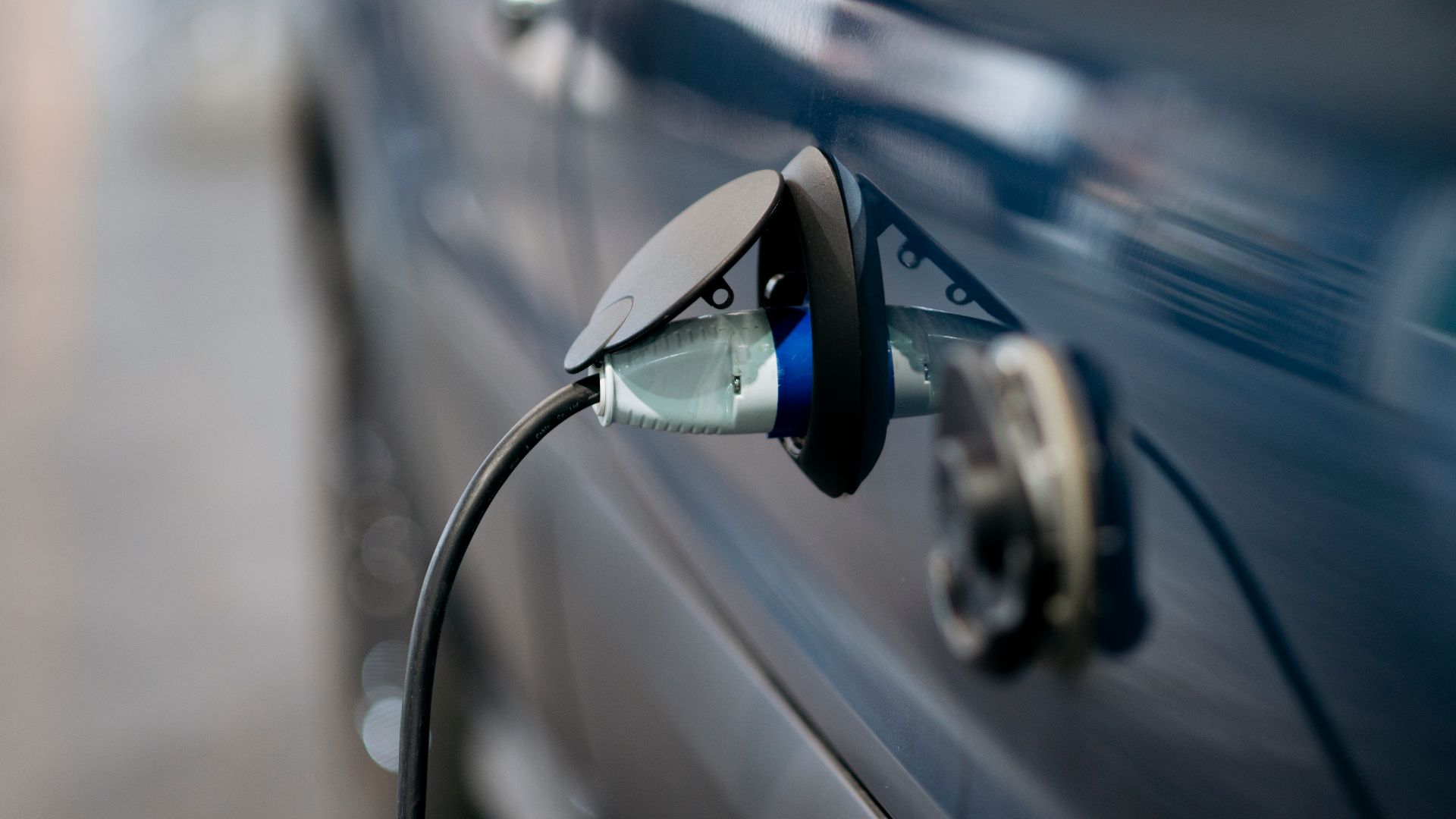 A blue electric car is at a charging station with the charging cable plugged into its port. The setting is urban and the time is daytime.