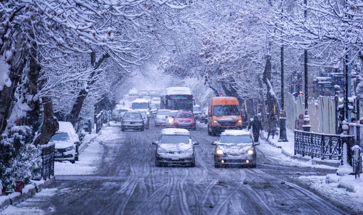 Heavy traffic on a snow-covered urban street lined with bare winter trees.