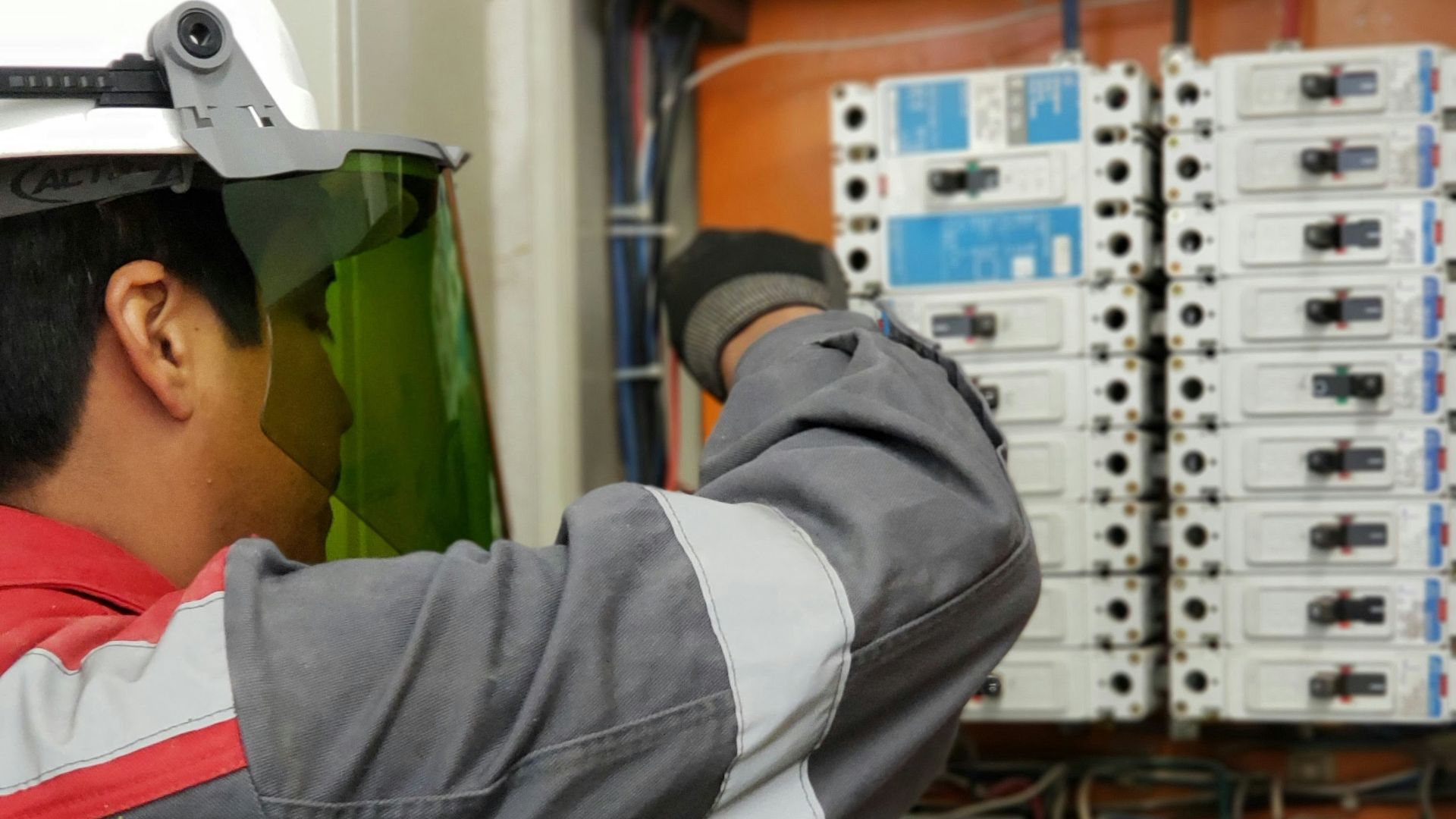 An electrician in a hard hat and face shield testing an electrical panel