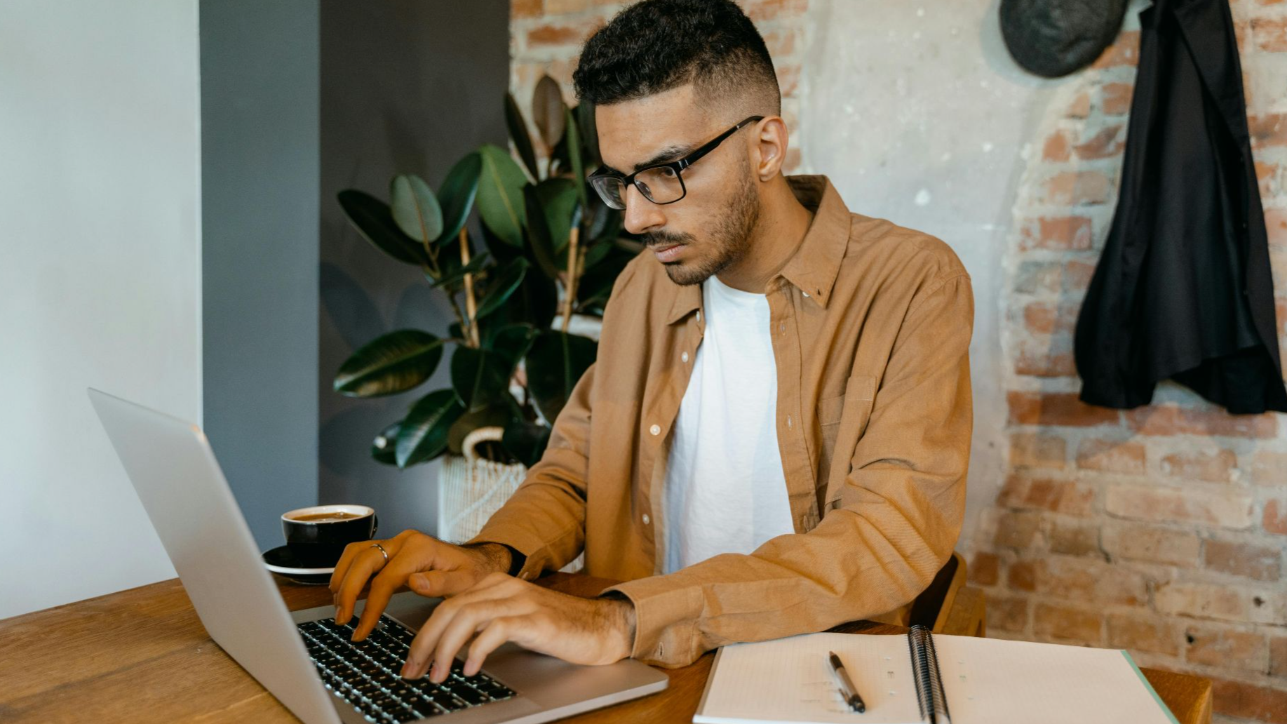 Man in casual office setting typing on laptop with coffee and notebook on desk.