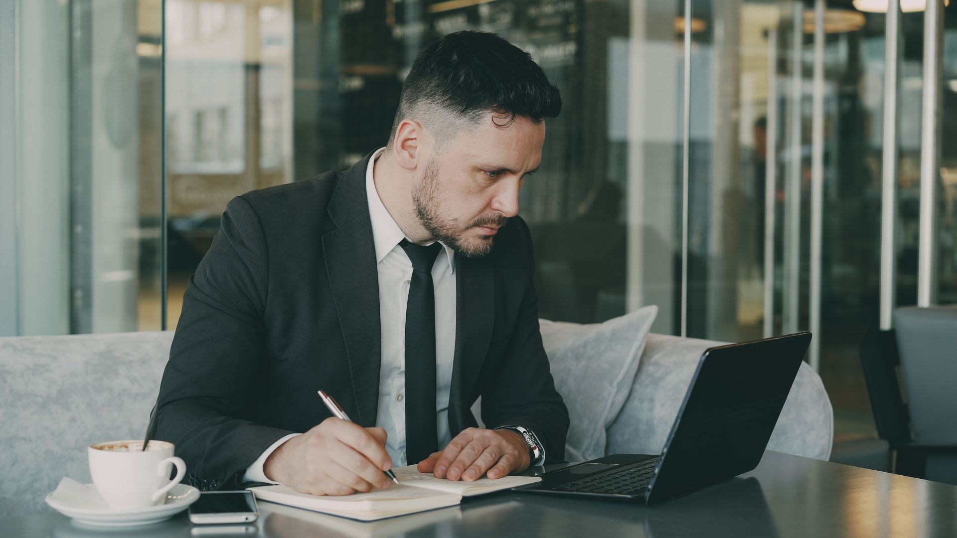A businessman in a suit working on a laptop and writing notes in a modern cafe.